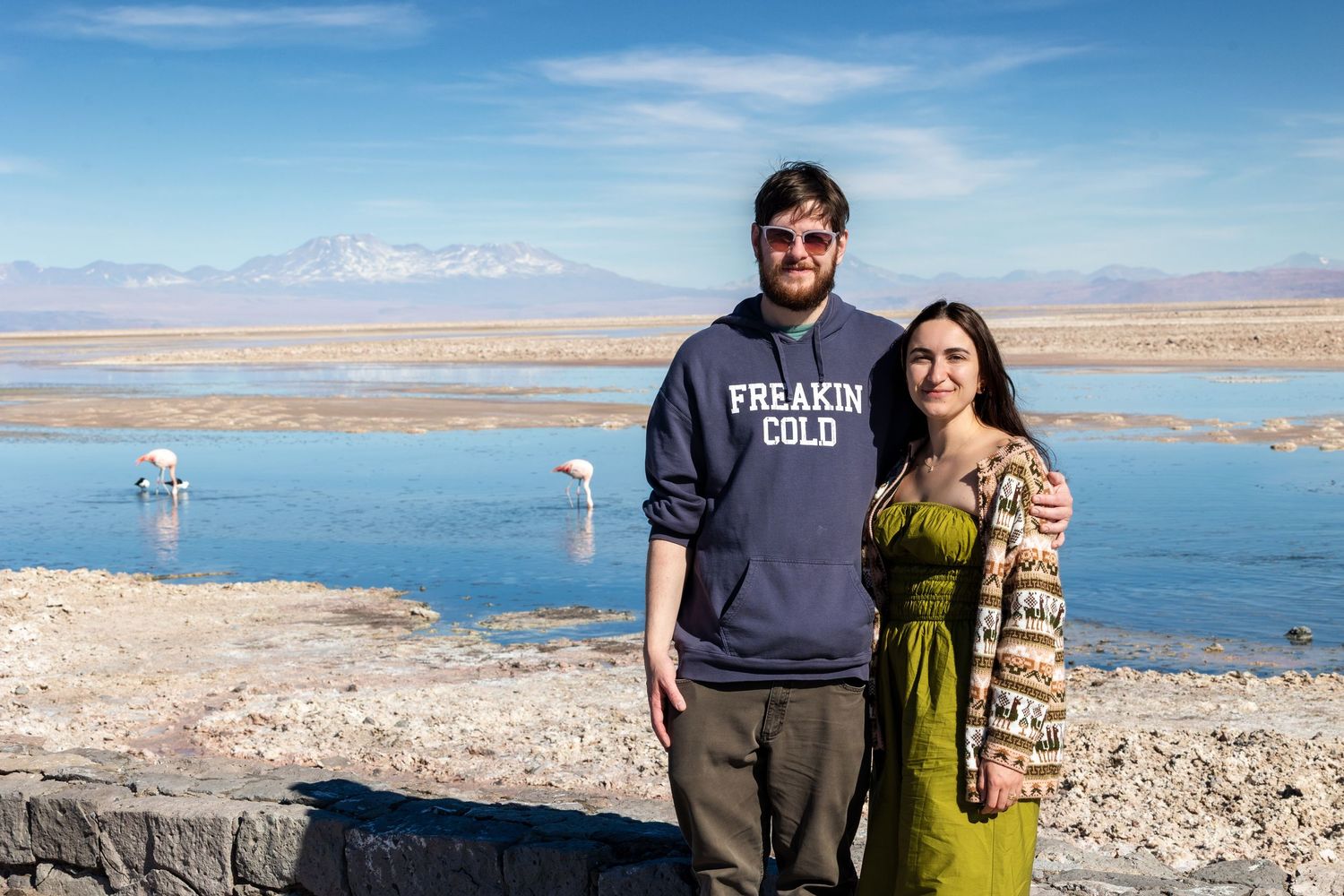Two people stand together on a rocky overlook with a bright blue lake and desert landscape in the background.