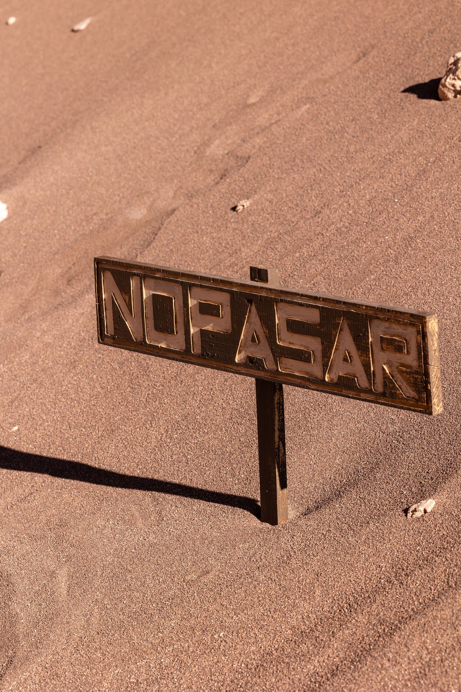 A weathered 'NO PASAR' (Do Not Pass) sign partially buried in desert sand with sepia-toned lighting.