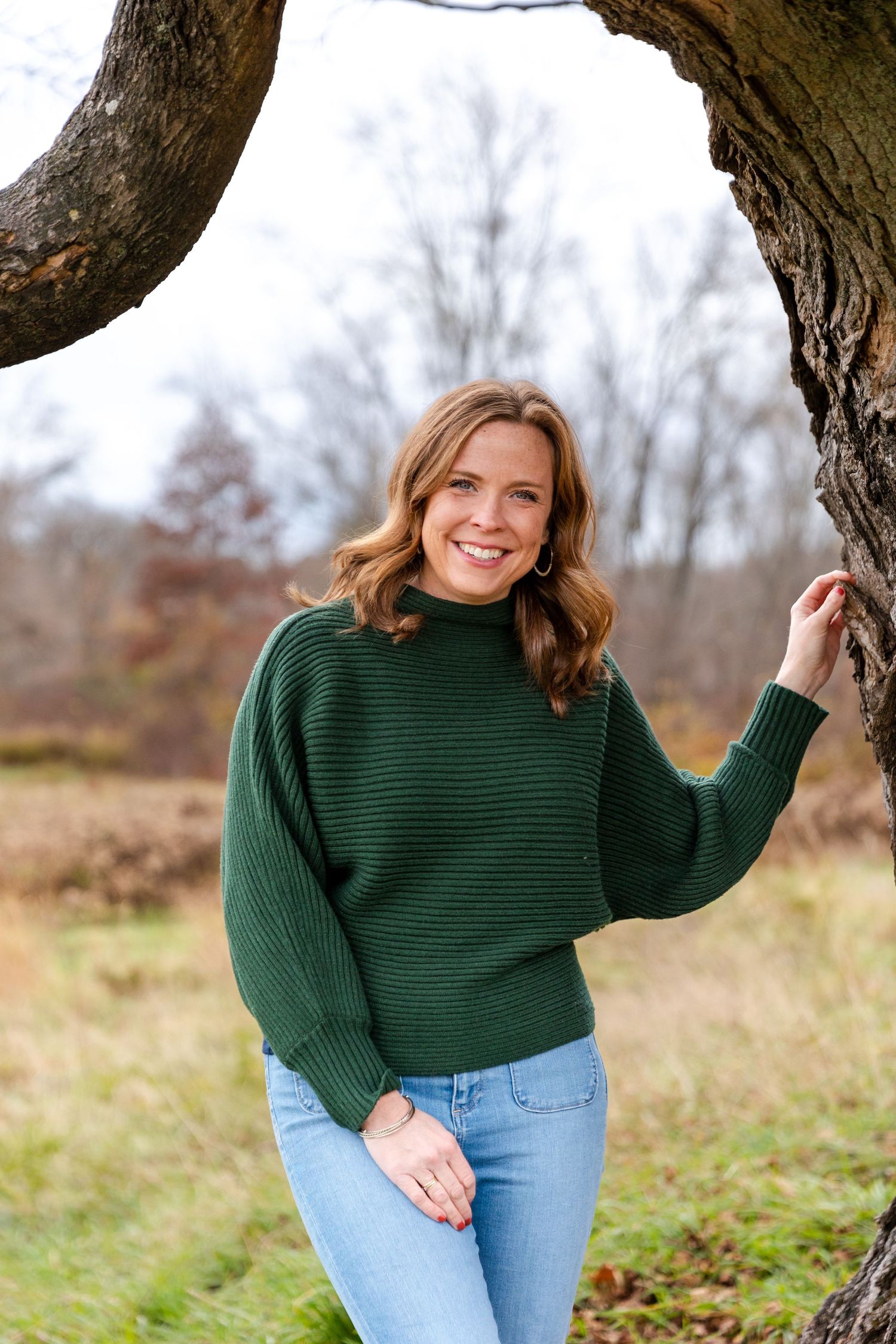 Person wearing ribbed green turtleneck sweater and jeans standing by tree in natural setting.