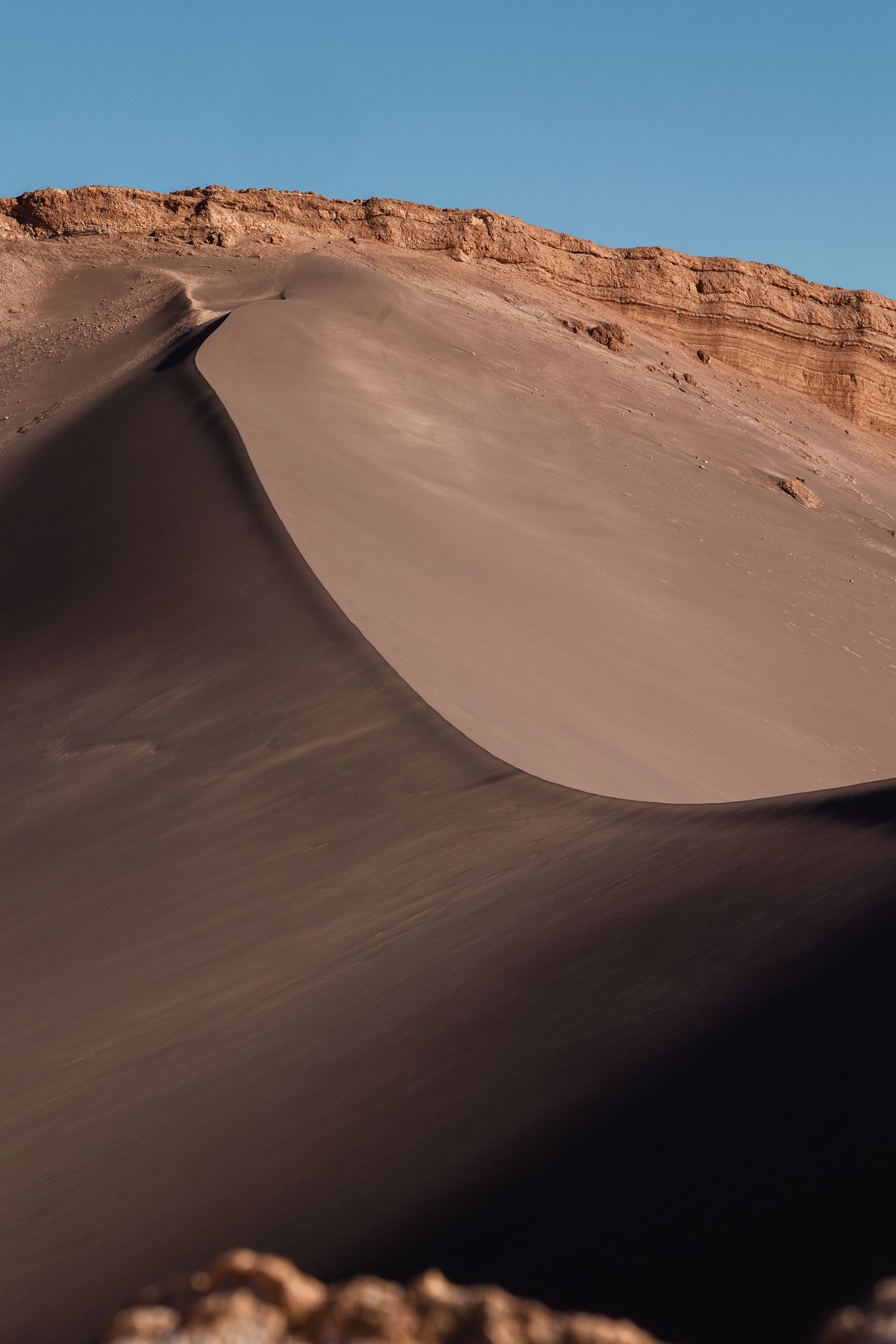 Smooth desert sand dune curves against orange rocky cliffs and bright blue sky in a dramatic desert landscape.