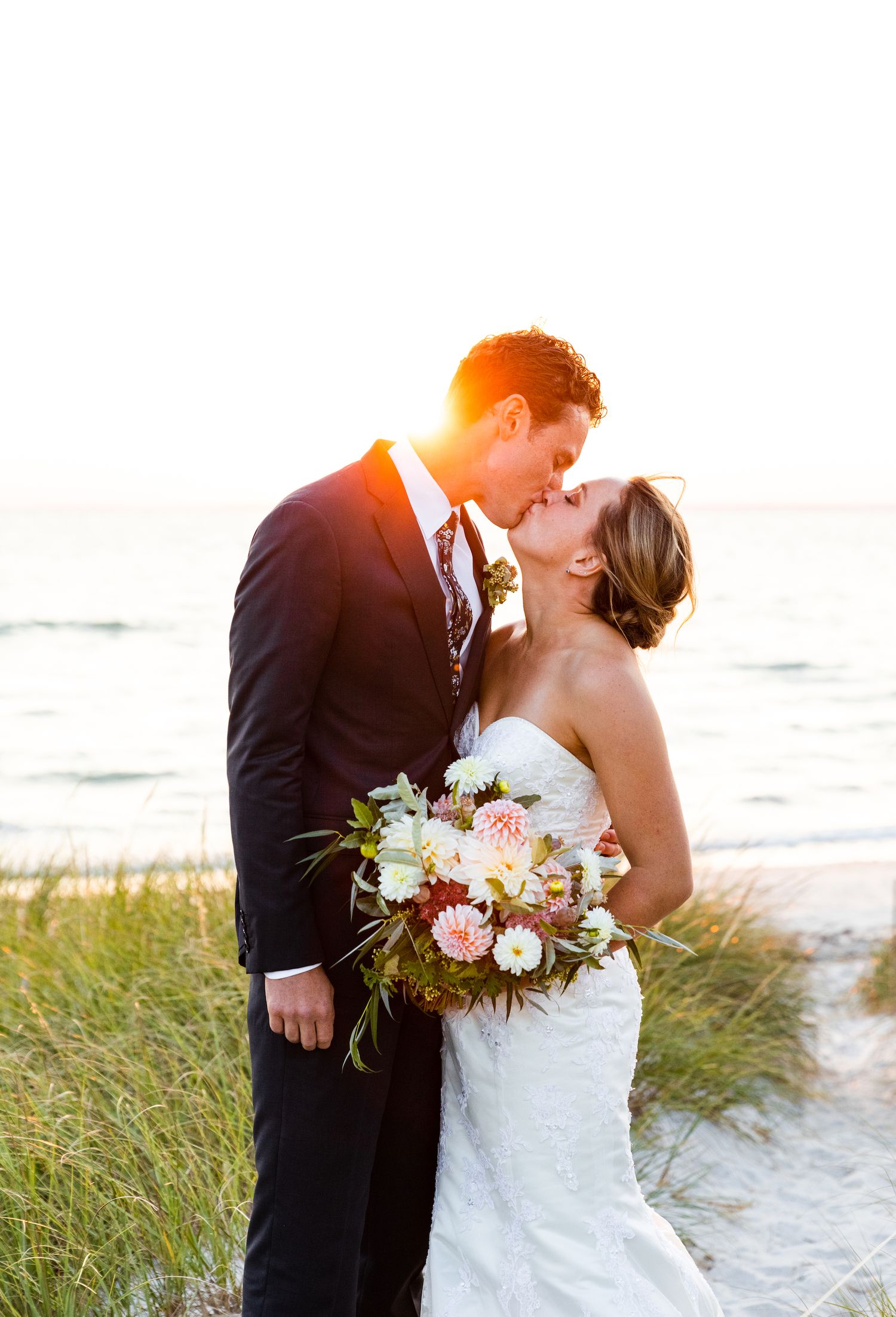 A romantic beach wedding photo of a couple kissing at sunset with a bridal bouquet.