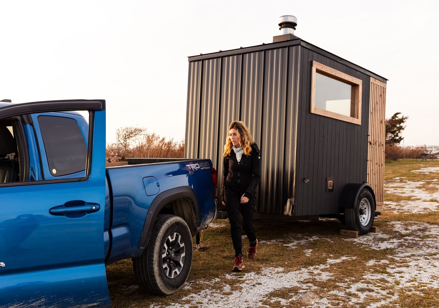 A blue pickup truck towing a small black modern mobile sauna trailer on snowy ground at sunset.