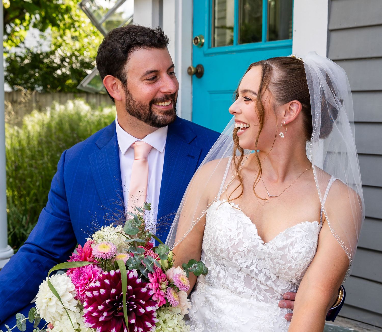 Two people in wedding attire share joyful laughter while posing outside a house with a bright turquoise door.