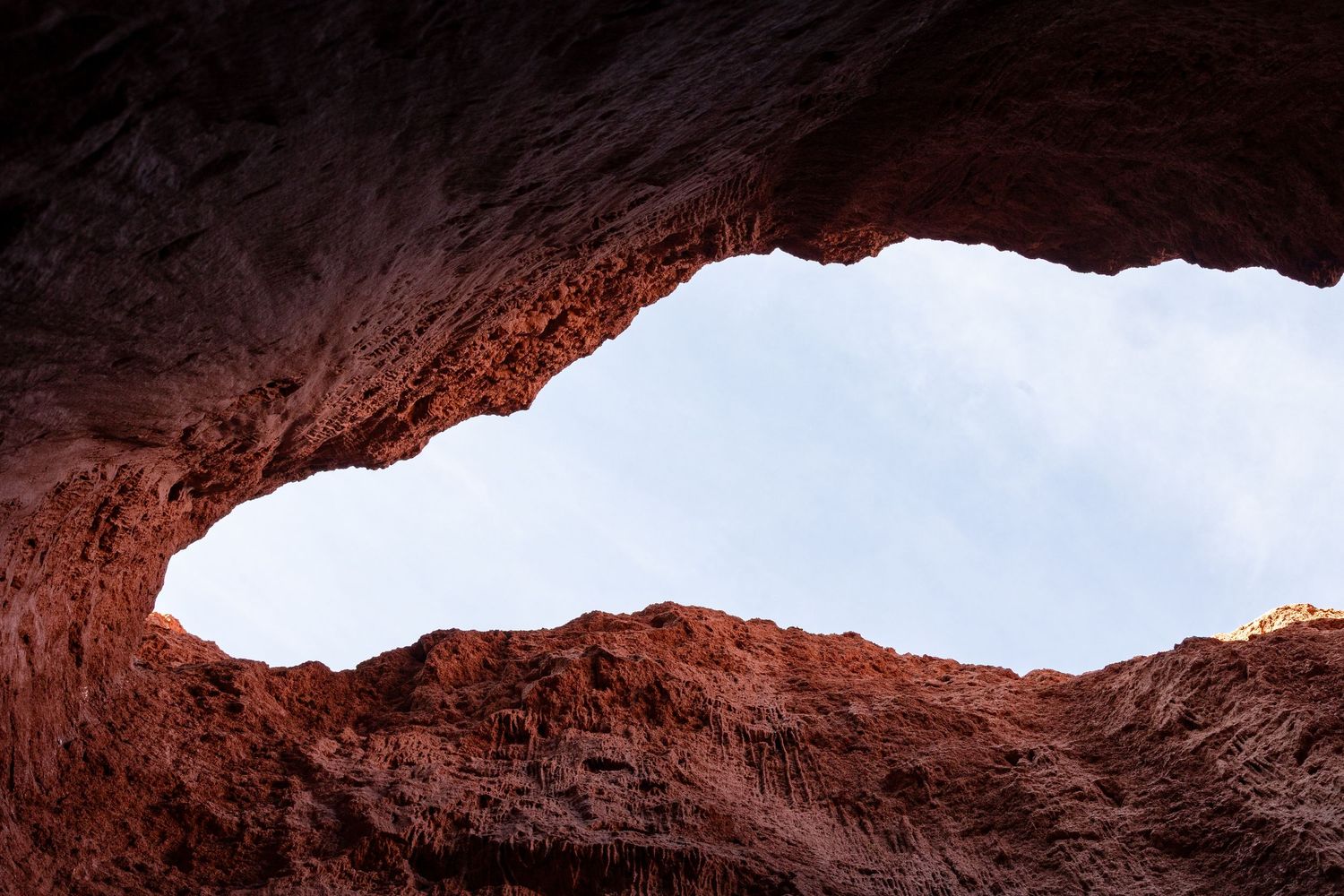 Looking up through a natural rock arch formation reveals bright sky framed by reddish sandstone walls.