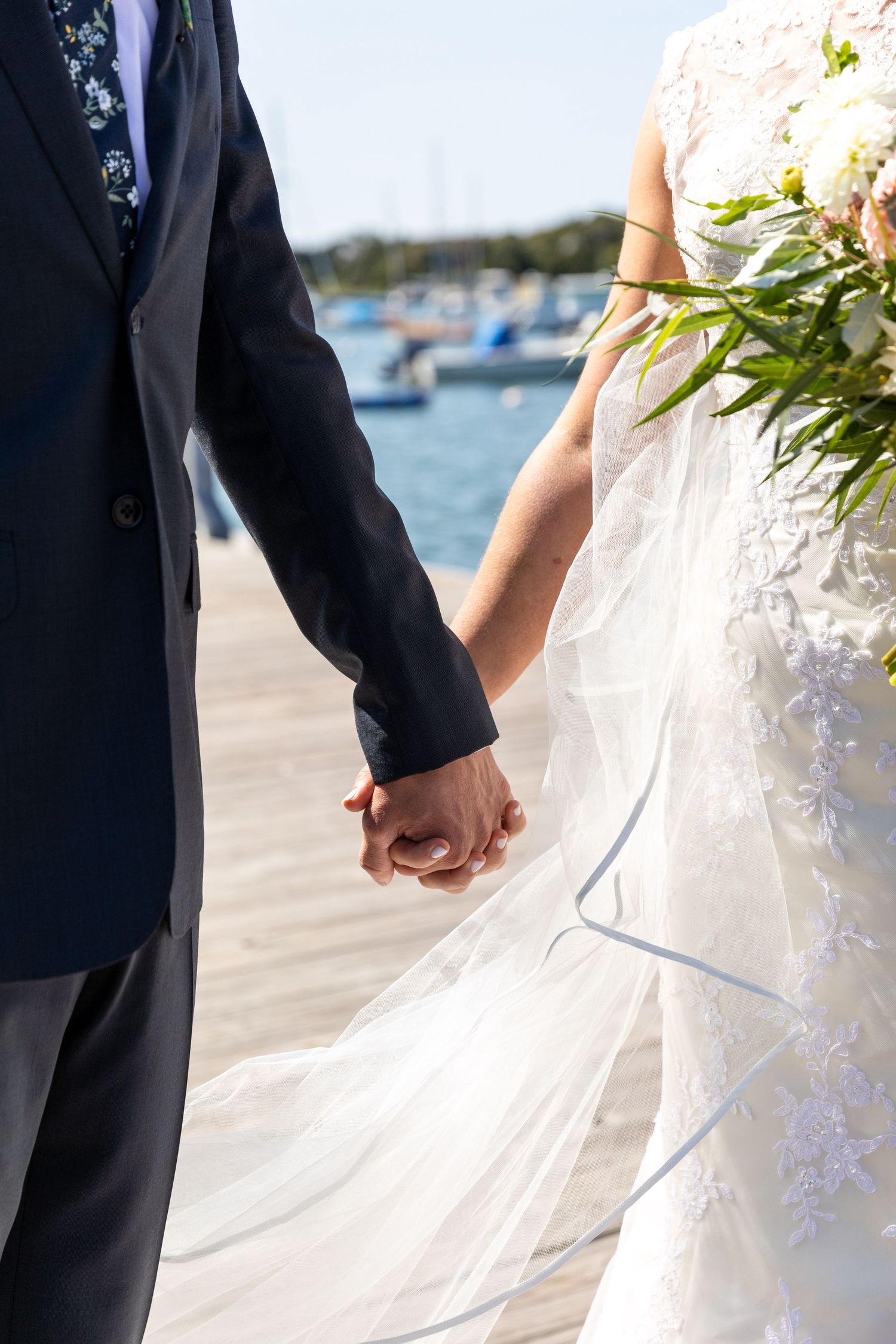 Newlywed couple holds hands while walking on a marina dock with boats in the background on their wedding day.