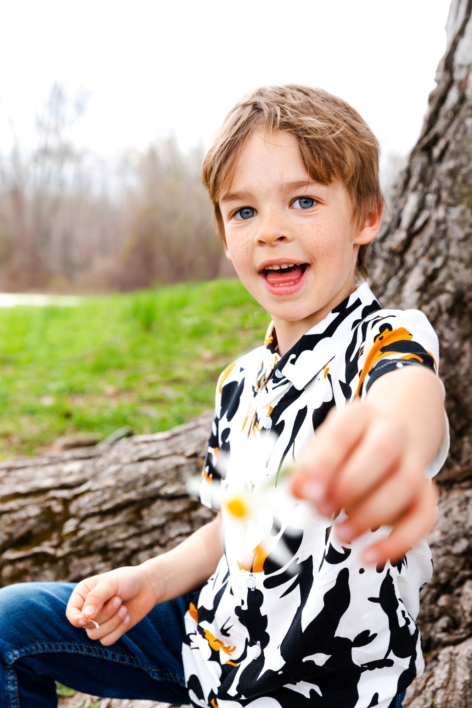 Energetic child in patterned shirt reaching towards camera while sitting by tree outdoors.