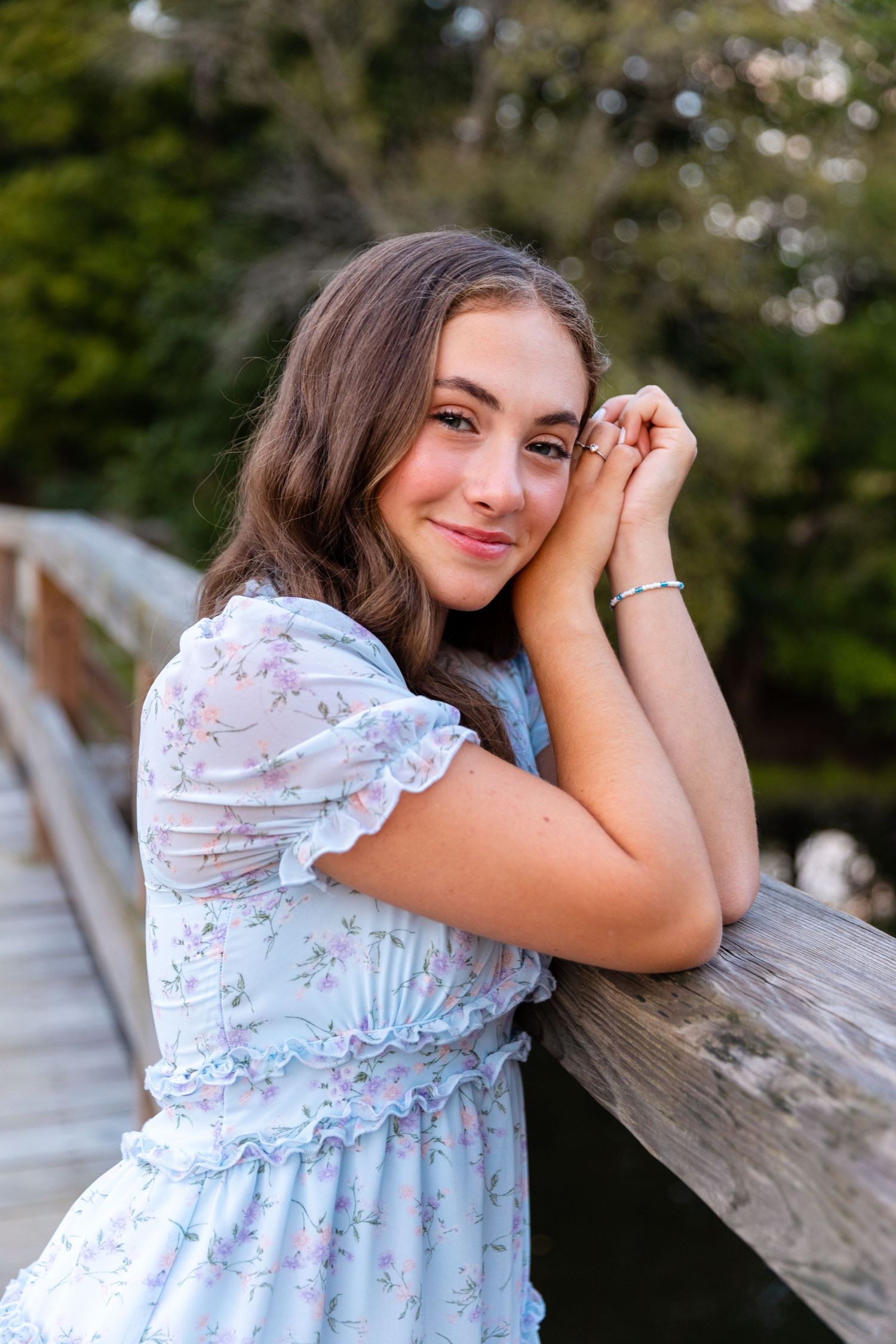 Figure in a floral dress leans against wooden railing during an outdoor portrait session.