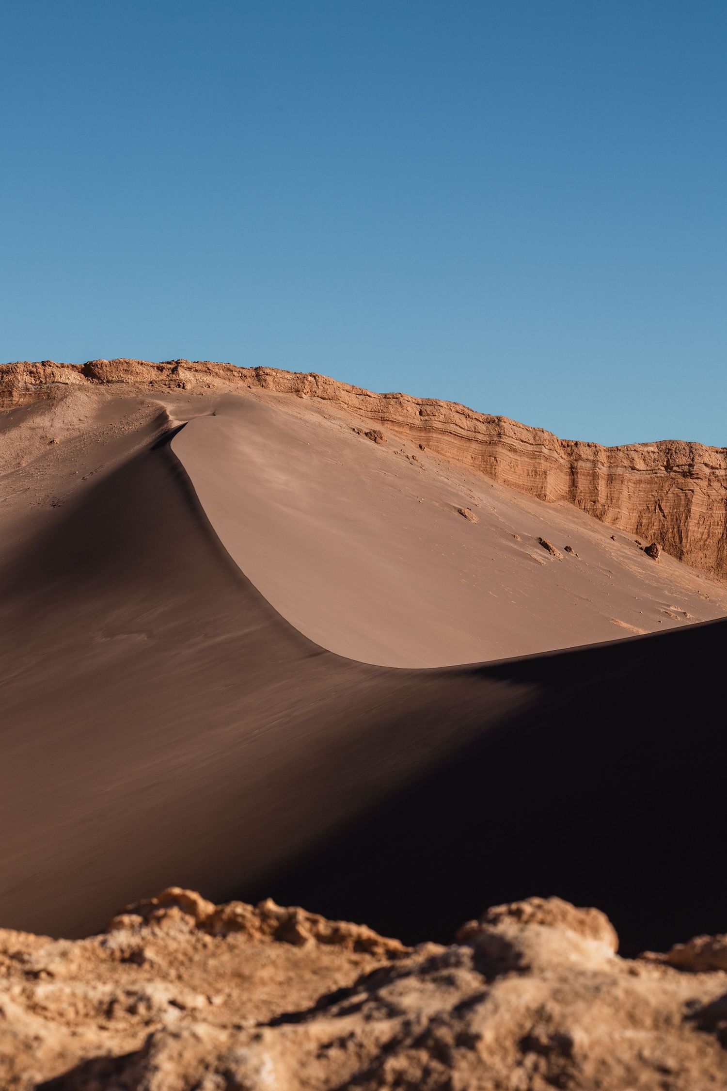 Smooth desert sand dunes rise against rocky cliffs and bright blue sky in an arid landscape.