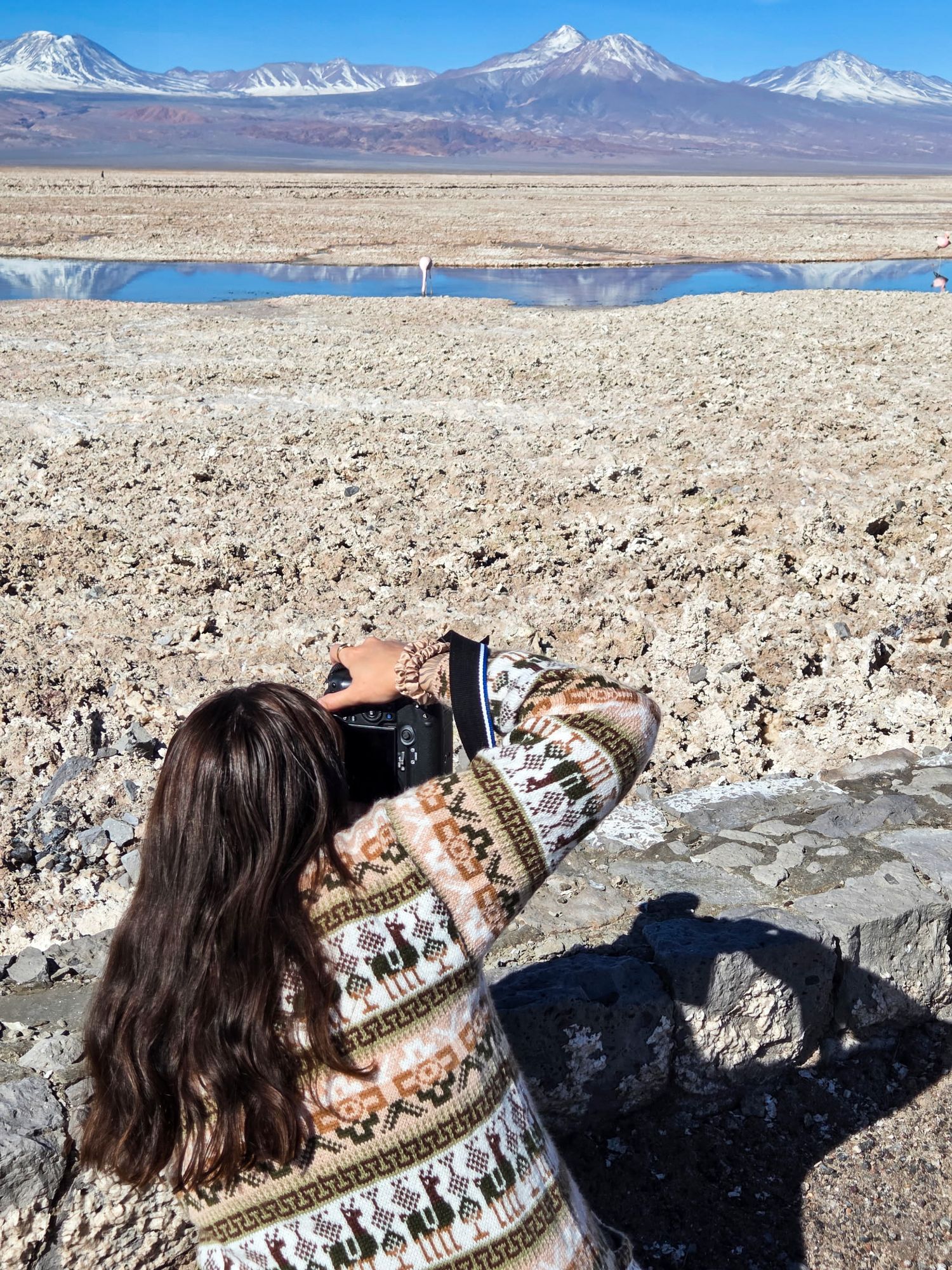 Photographer in patterned sweater captures snowy mountain landscape reflected in desert salt flats.