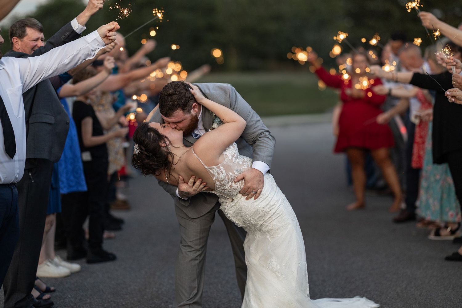 Wedding couple shares romantic dip kiss while guests toss confetti during sparkler exit celebration at dusk.