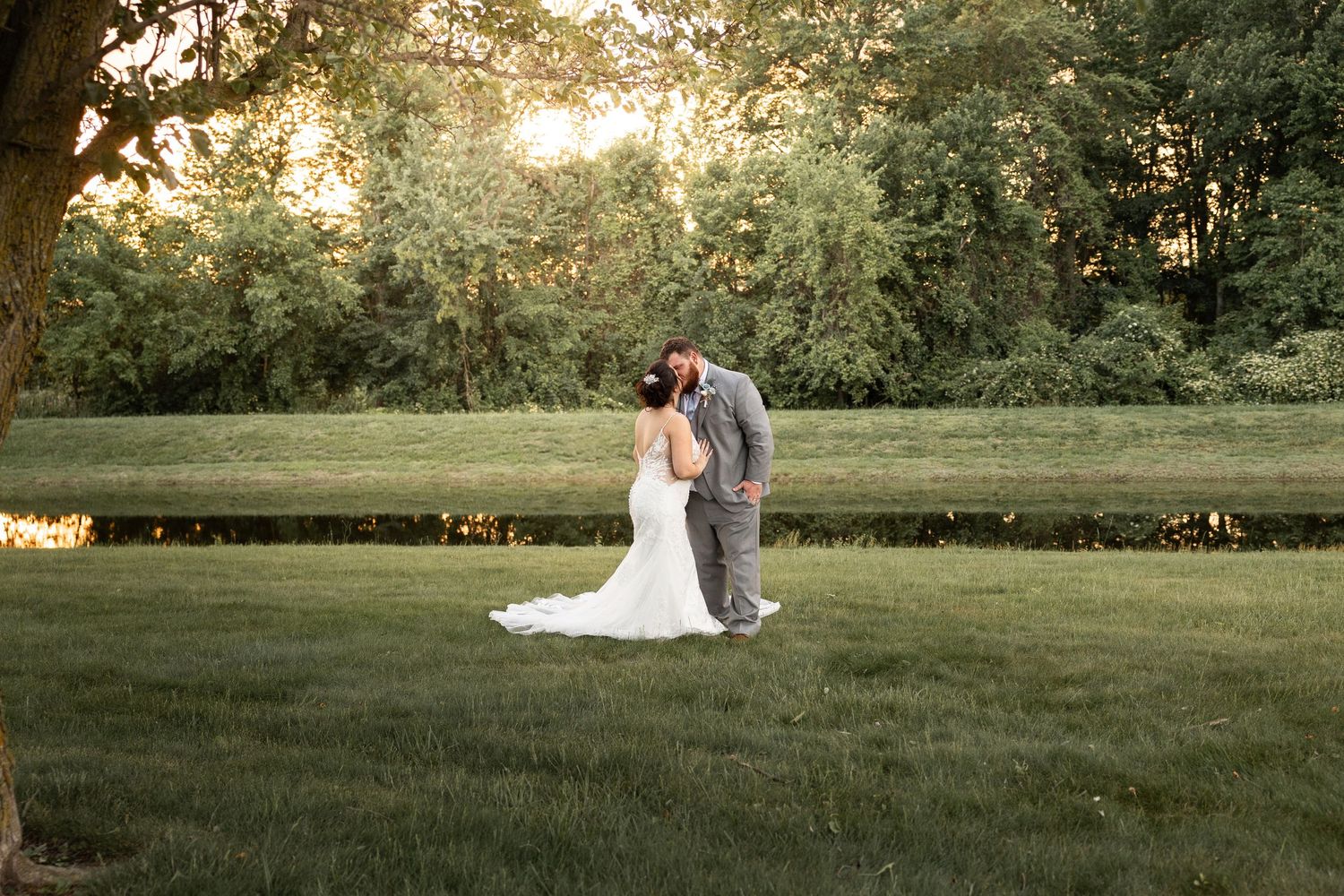 Two people share a romantic moment at sunset on a lush green lawn with trees and golden light in the background.