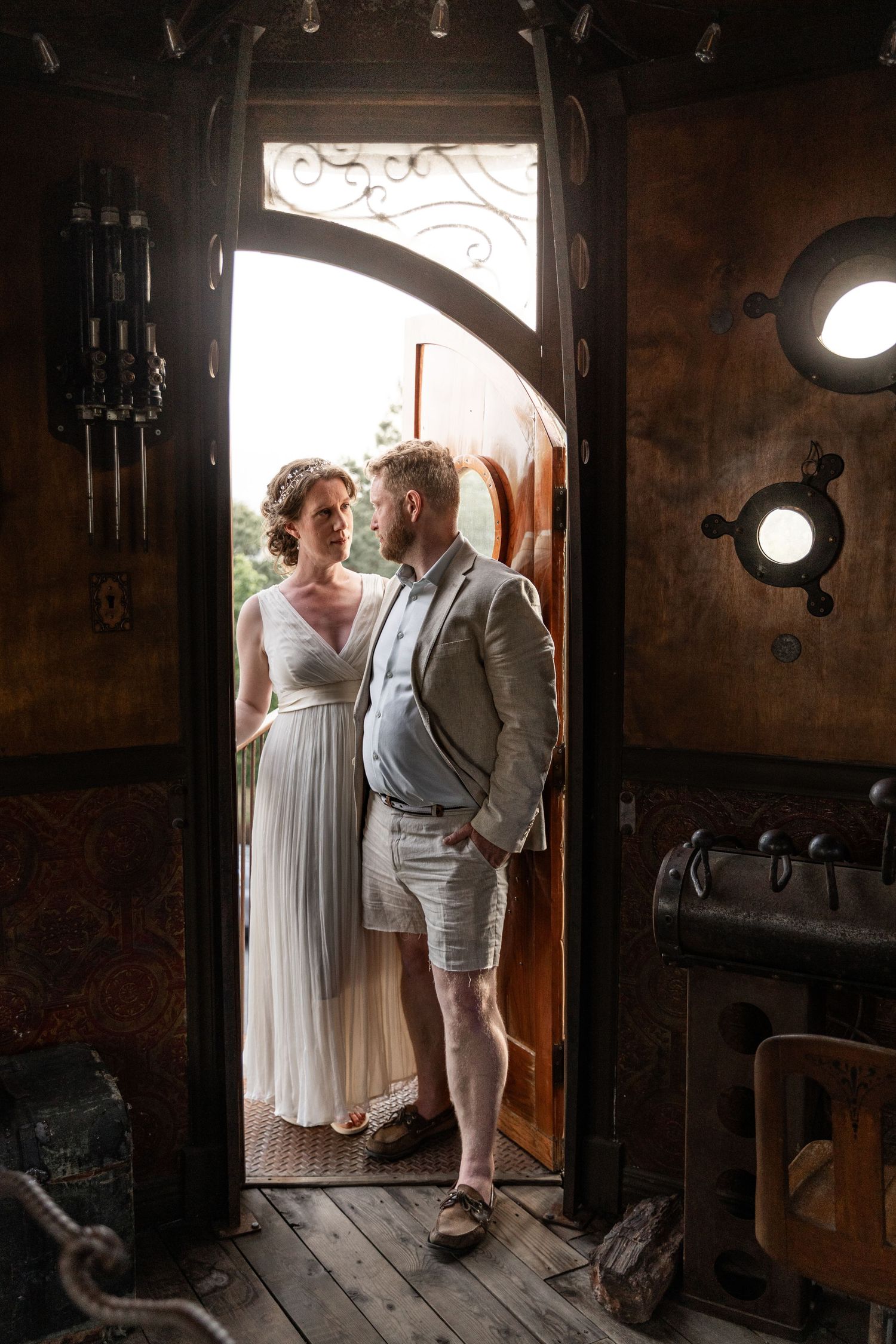 A couple in elegant wedding attire stands in the doorway of a vintage wooden boat interior with porthole windows.
