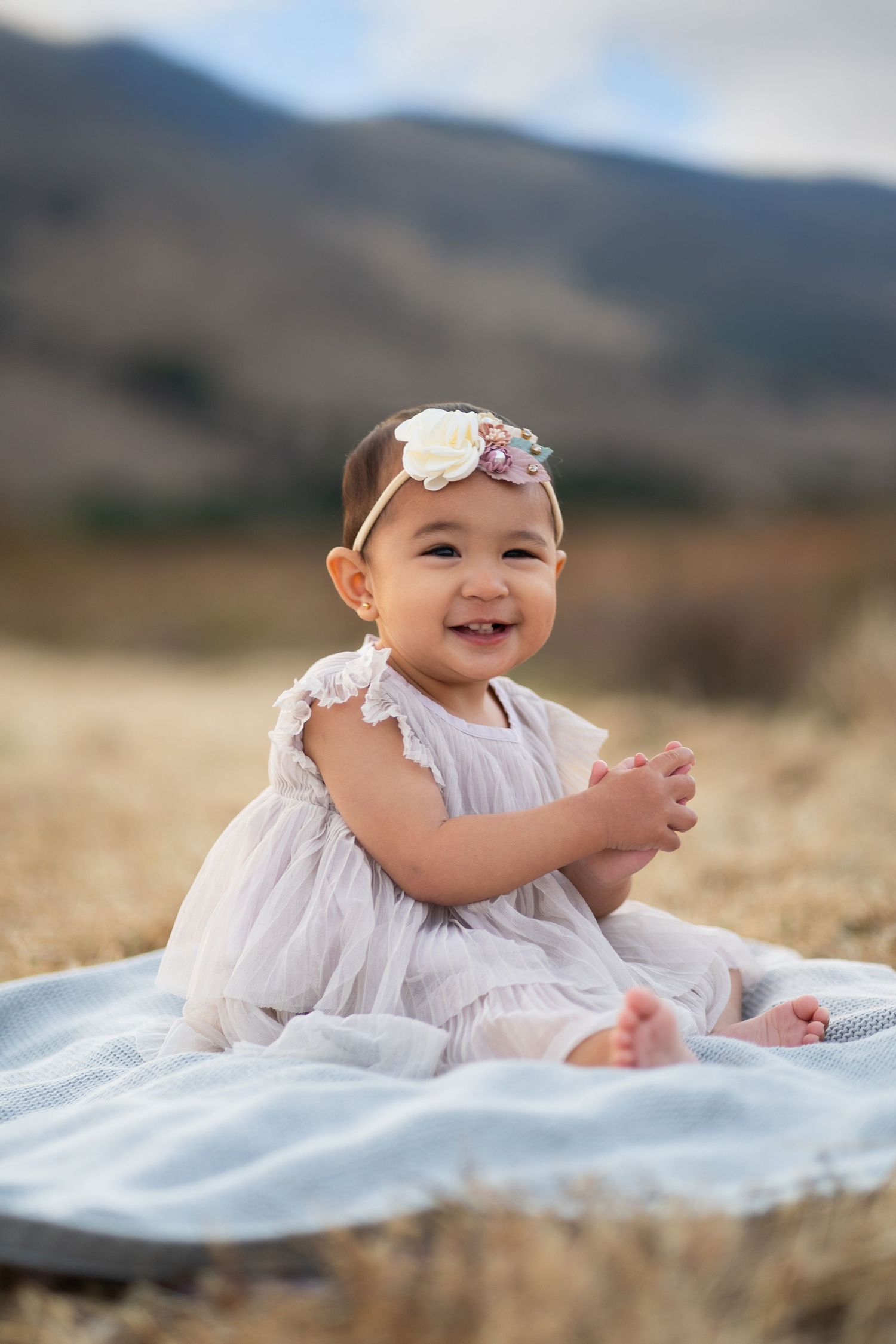 Baby in white dress and floral headband sits on blue blanket in mountain field.