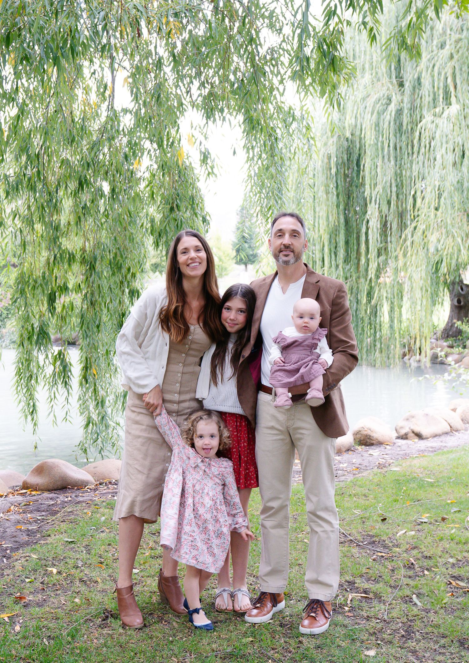 Family of five poses together under weeping willow trees in coordinated neutral outfits.