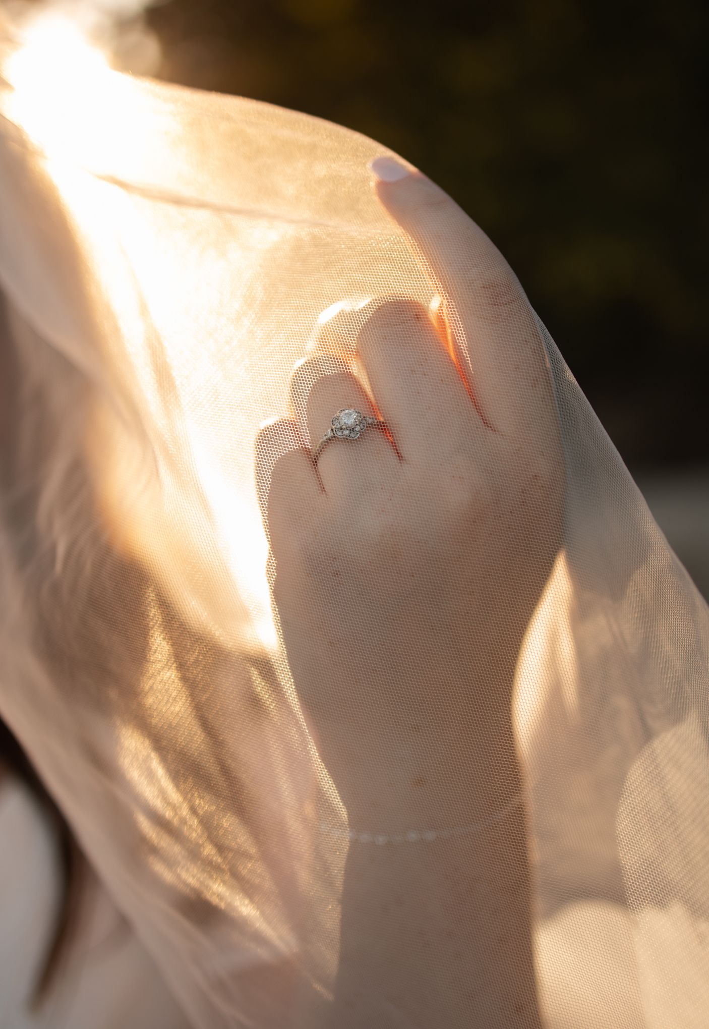 Sunlit engagement ring photographed through delicate white veil fabric.