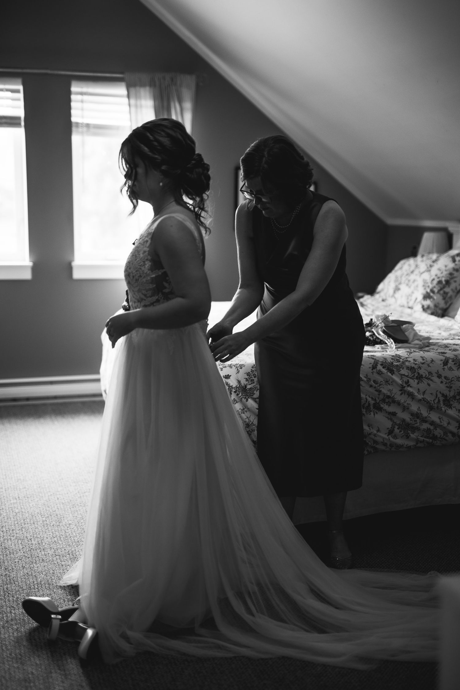 A black and white intimate moment showing final wedding dress preparations in an upstairs bedroom.