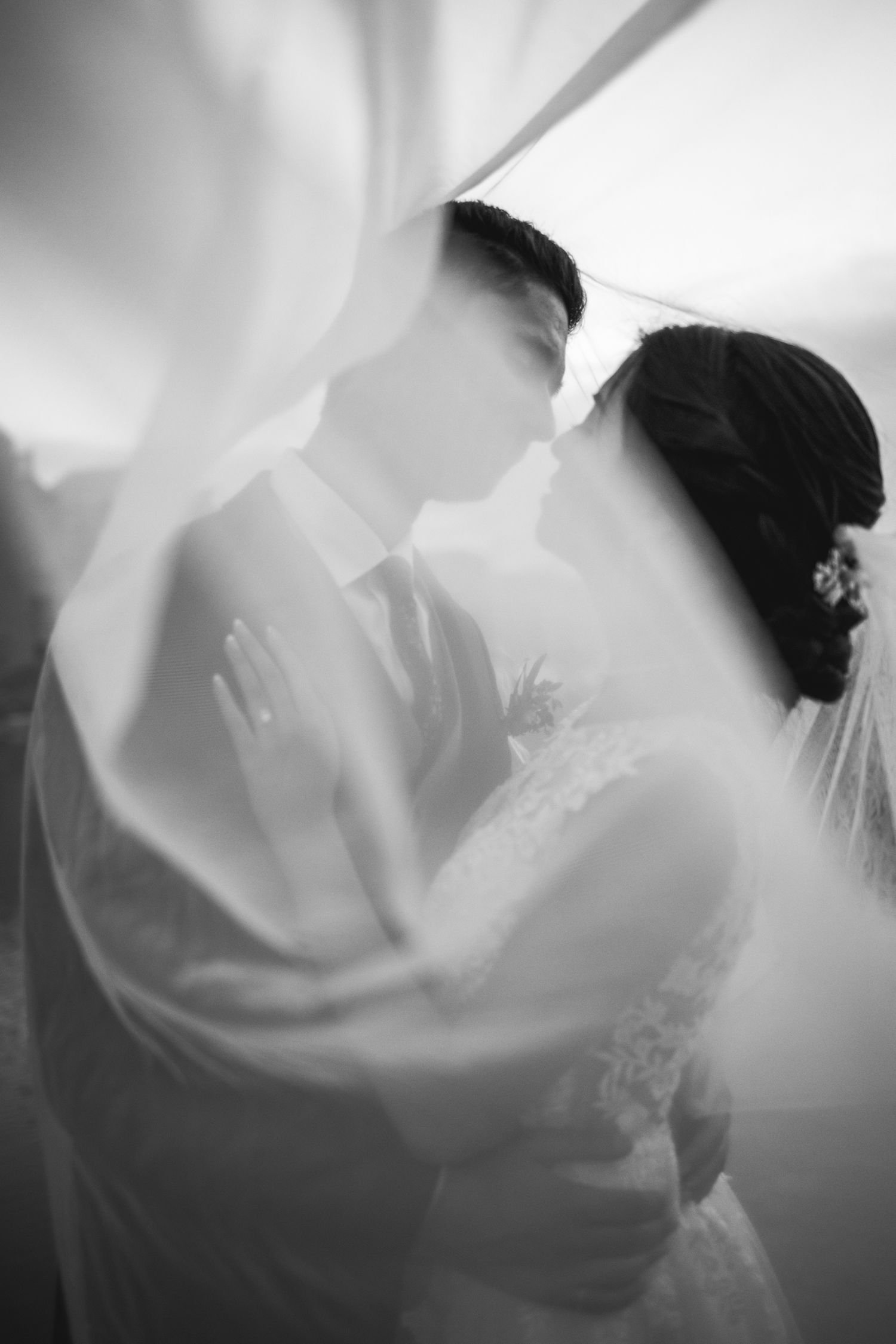 An ethereal wedding moment captured through flowing veil fabric in black and white.