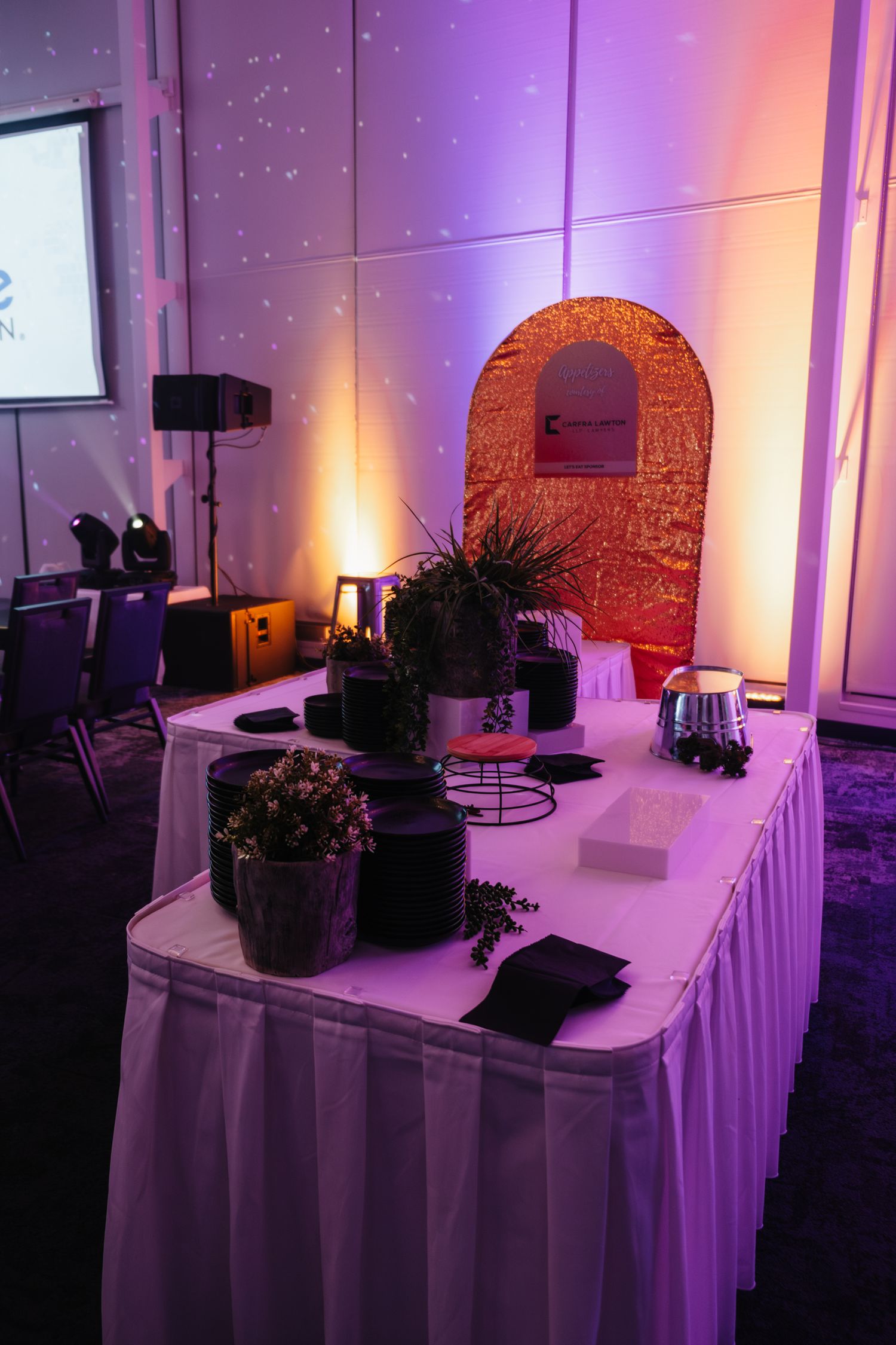 Event setup with decorative arch and potted plants under purple and orange ambient lighting.