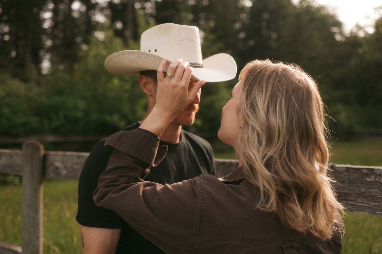 A couple embraces outdoors as woman reaches up to lift cowboy hat from person wearing brown shirt at sunset.