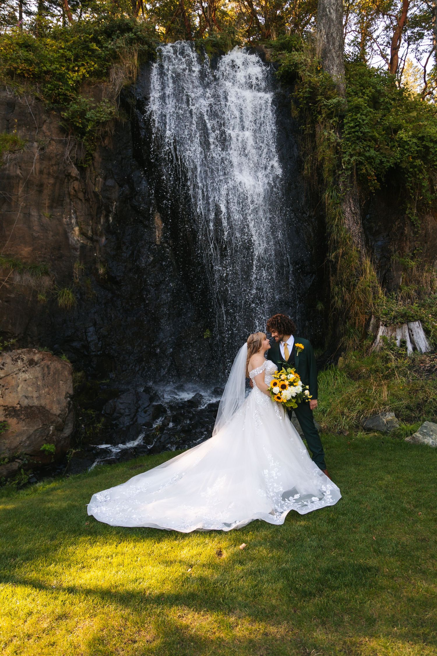 A couple embraces in front of a majestic waterfall on their wedding day, with sunlight streaming across green grass.