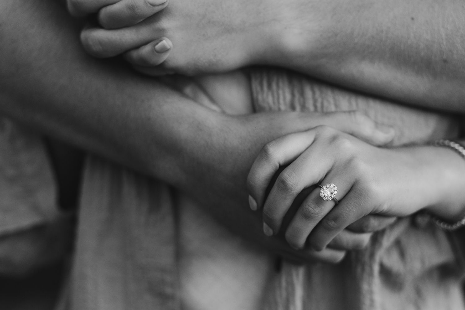 A black and white close-up photograph of intertwined hands with an engagement ring.