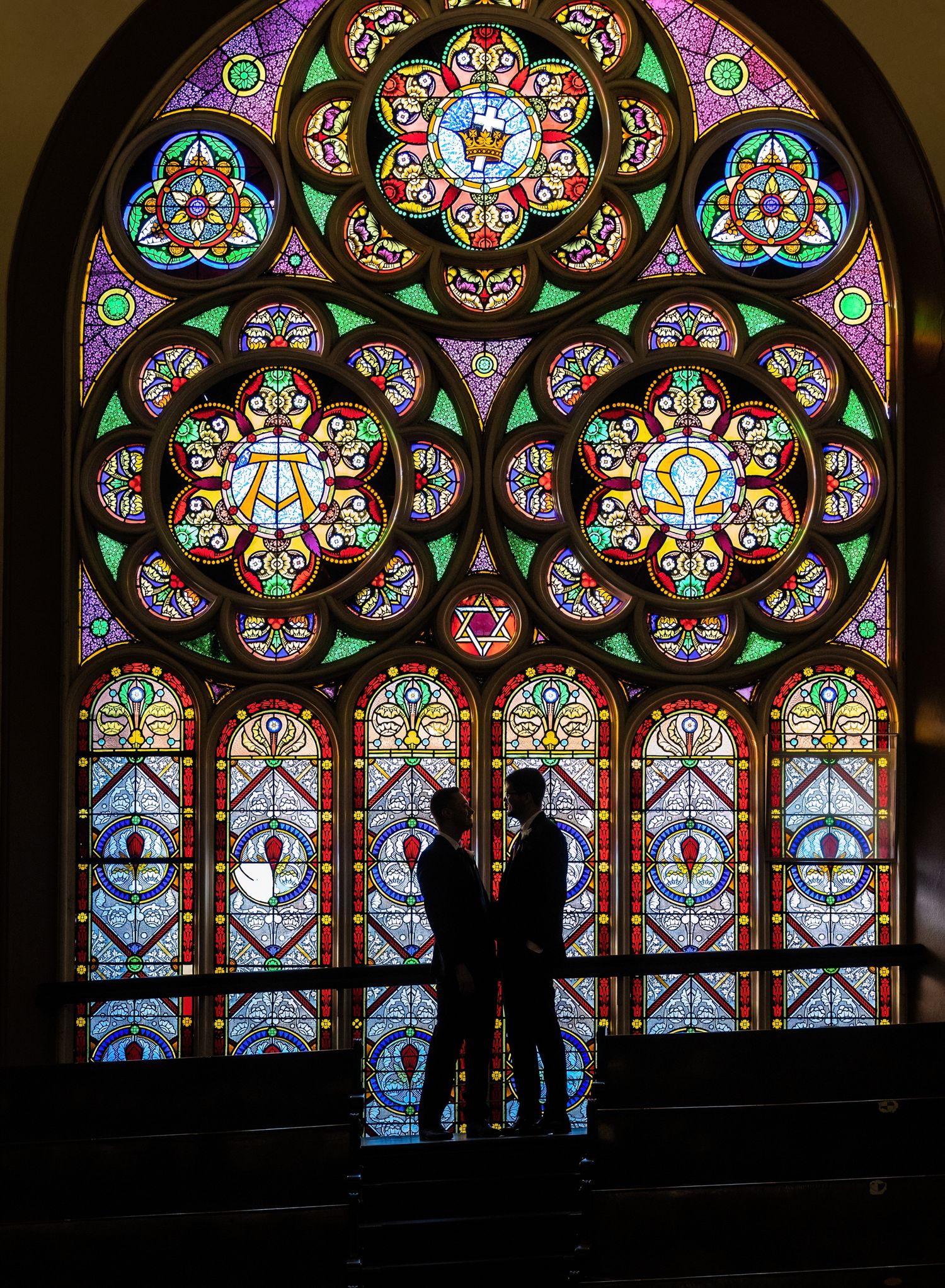 Two silhouettes stand before a magnificent stained glass cathedral window with intricate colorful patterns.