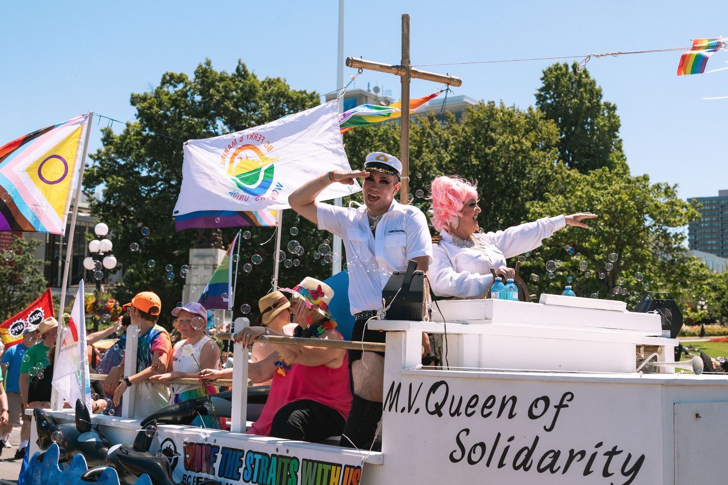 Pride parade participants wave from a vehicle decorated with flags and banners during a sunny celebration.