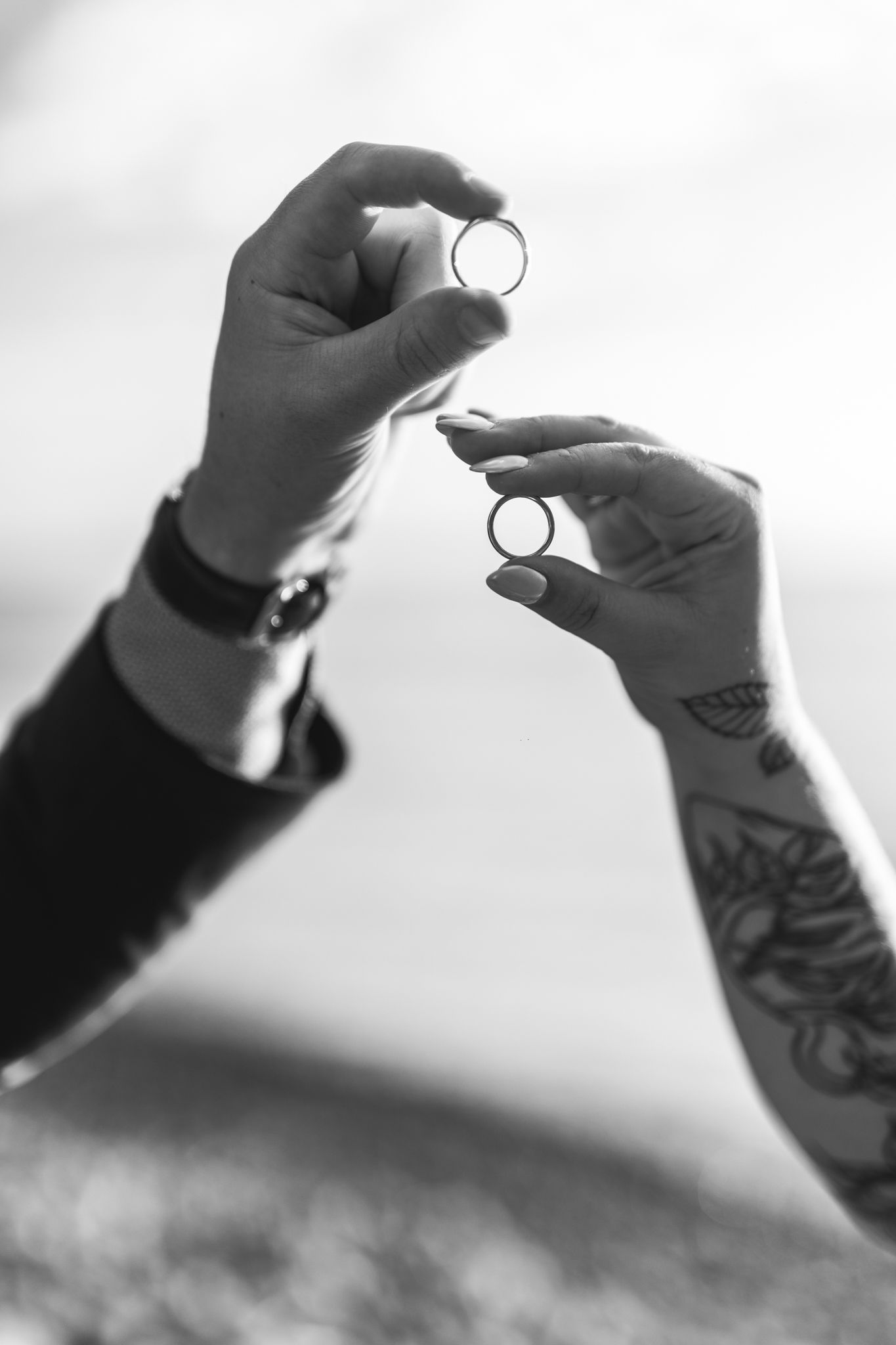 Black and white photo of hands holding wedding rings with one arm showing artistic tattoo details.