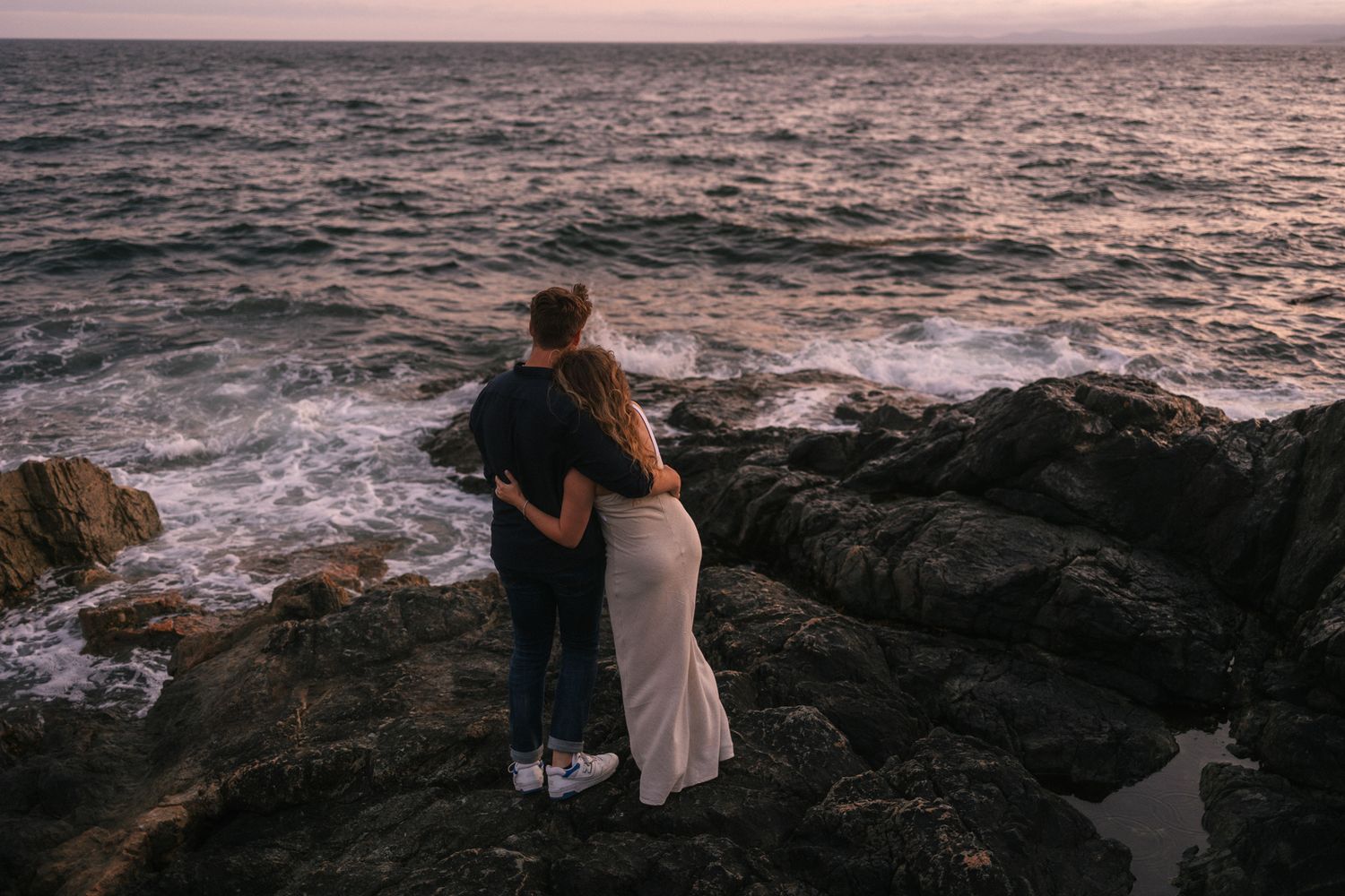 A romantic couple watches the sunset from a rocky coastal viewpoint overlooking dramatic ocean waves.