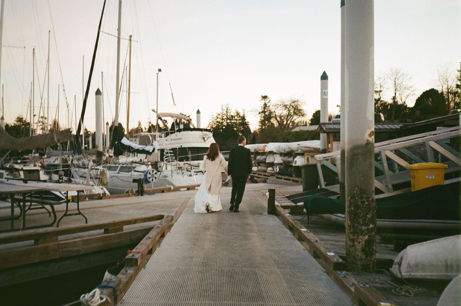 Figures walking along a marina dock at sunset with sailboats and yachts moored on either side.