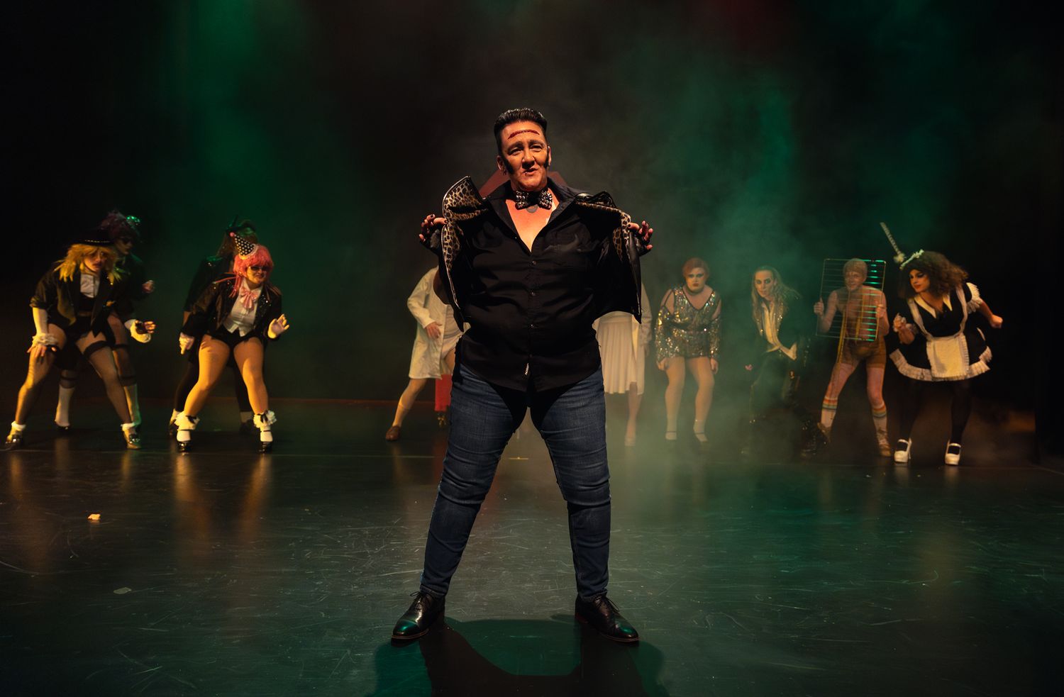Performer in black leather jacket strikes dramatic pose on dark stage with backup dancers silhouetted behind in green fog.