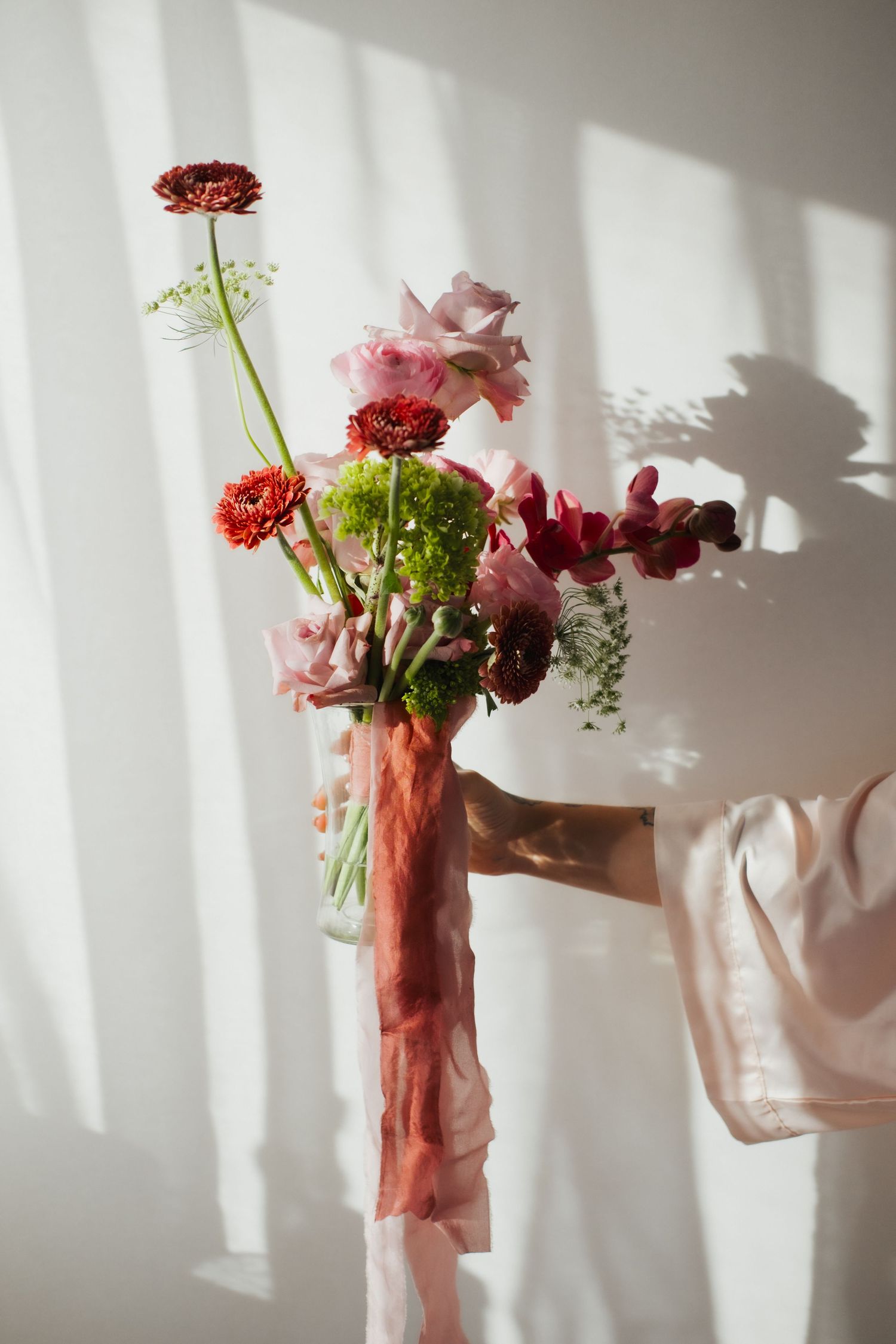 Pink and red flowers arranged in a glass vase casting dramatic shadows on white wall.