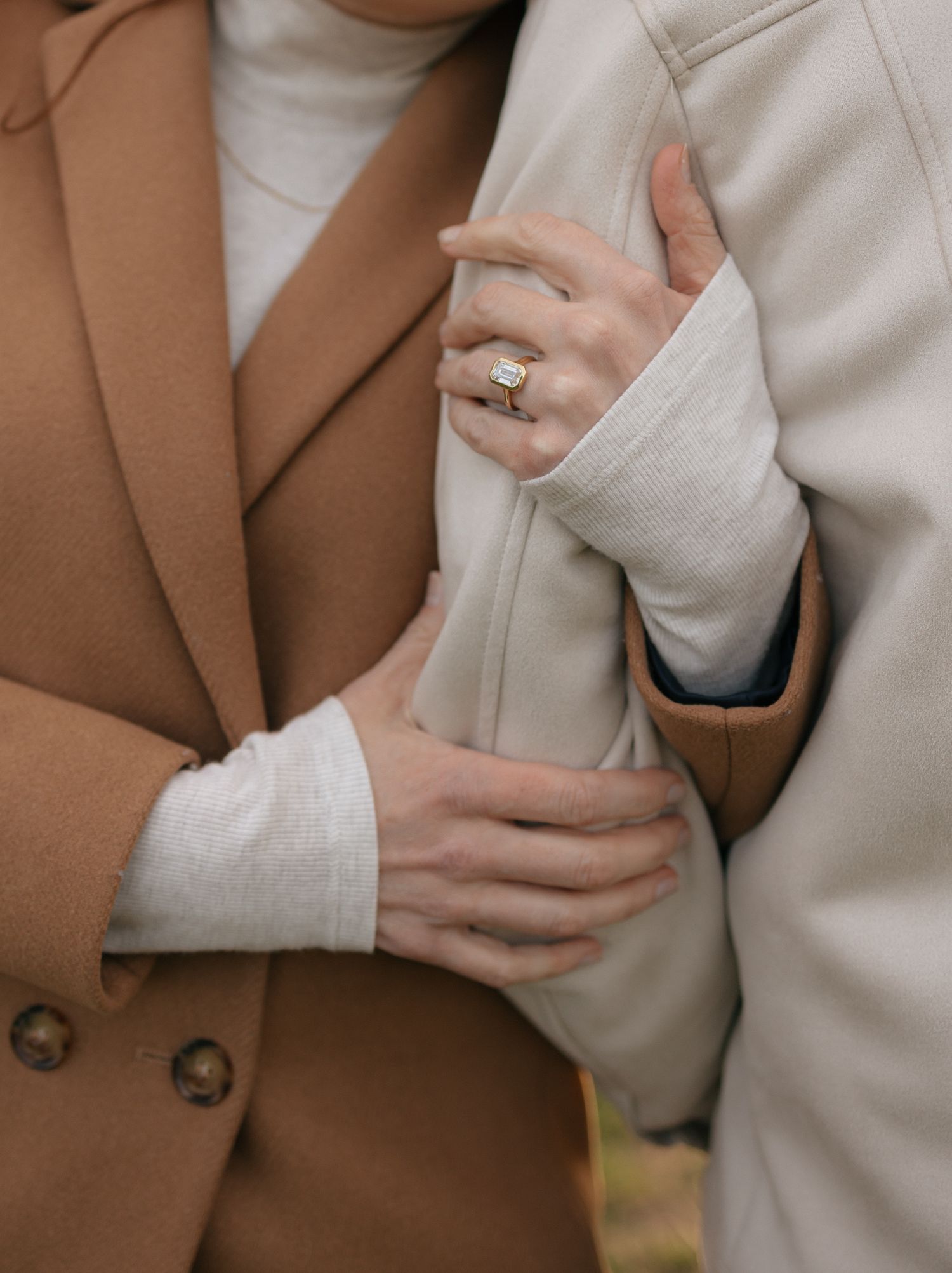 Close-up shot of hands wearing brown and beige coats with one hand displaying an elegant ring on their finger.