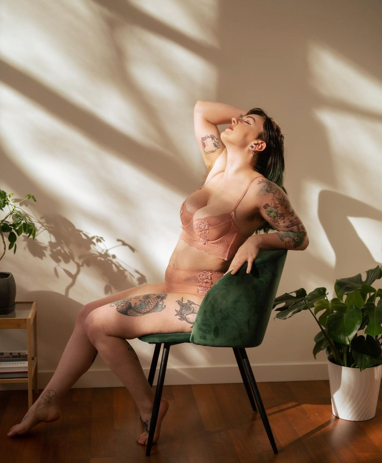 Person in pink lingerie posing on green velvet chair with sunlight and plants in background.