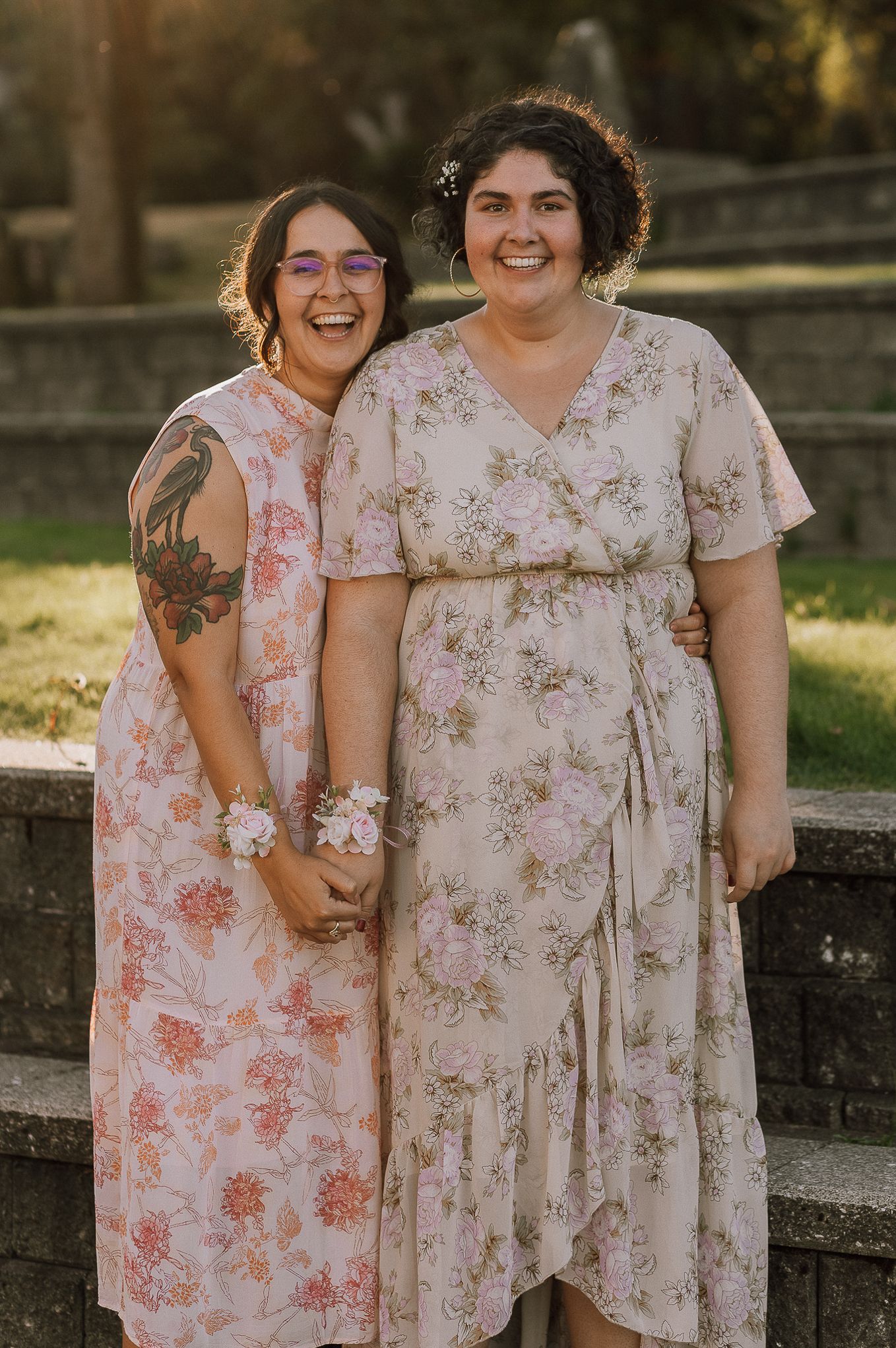 Two wedding guests in floral dresses pose together outdoors during golden hour.