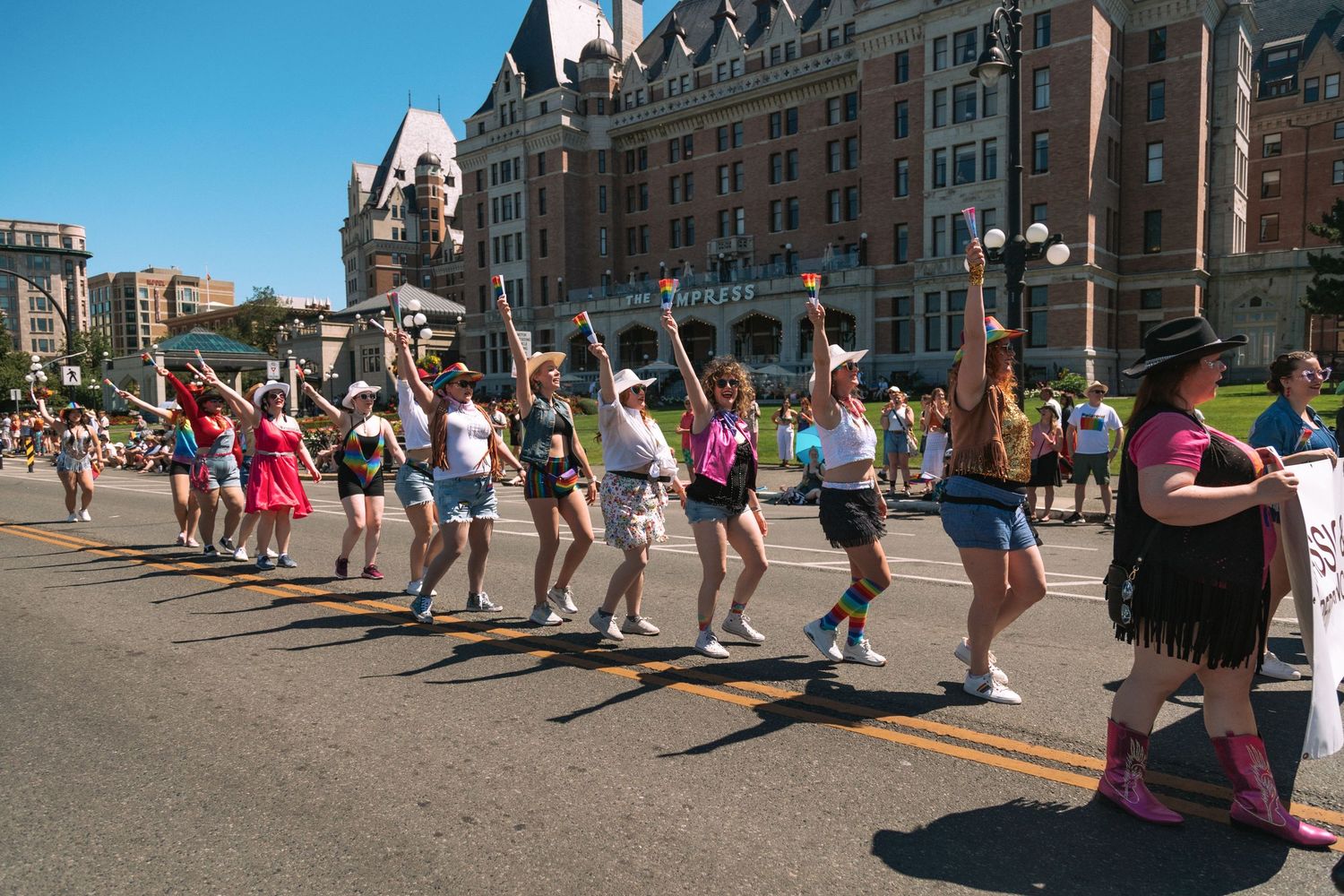 A line of cheerleaders perform synchronized jumps and dance moves during an outdoor parade event in a sunny cityscape.
