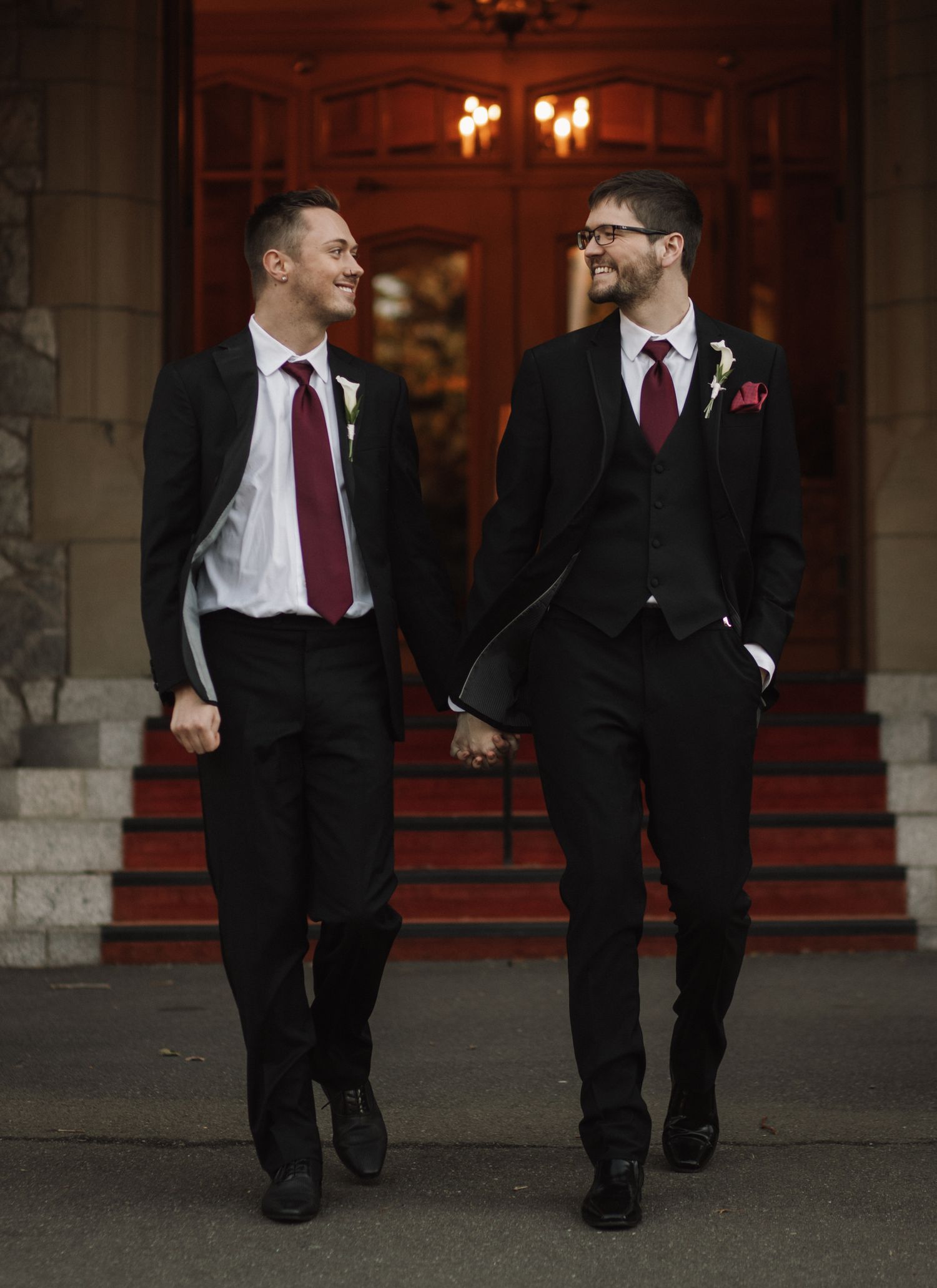 Two wedding guests in black suits and burgundy ties share a happy moment in an archway with warm lighting.