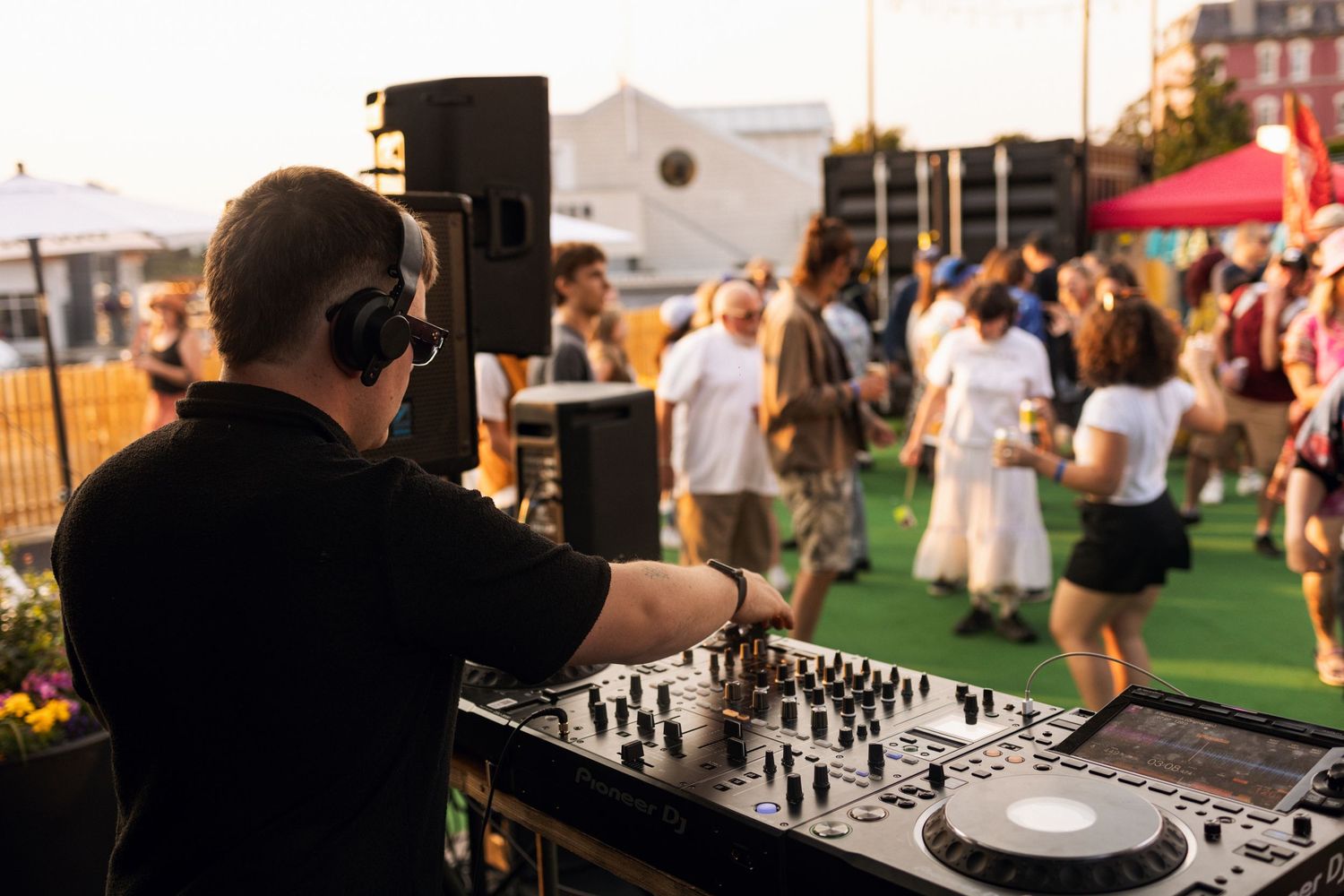 A DJ performs at an outdoor event with mixing equipment while people gather on artificial turf in the background.