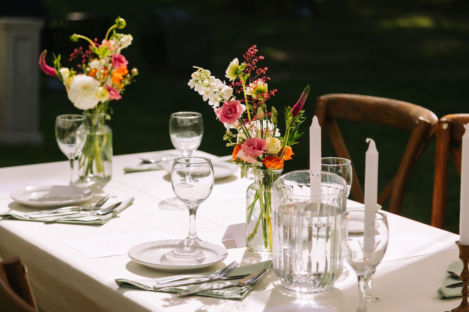 An elegant table setting with small glass vases of colorful flowers, wine glasses, and place settings on white linens.