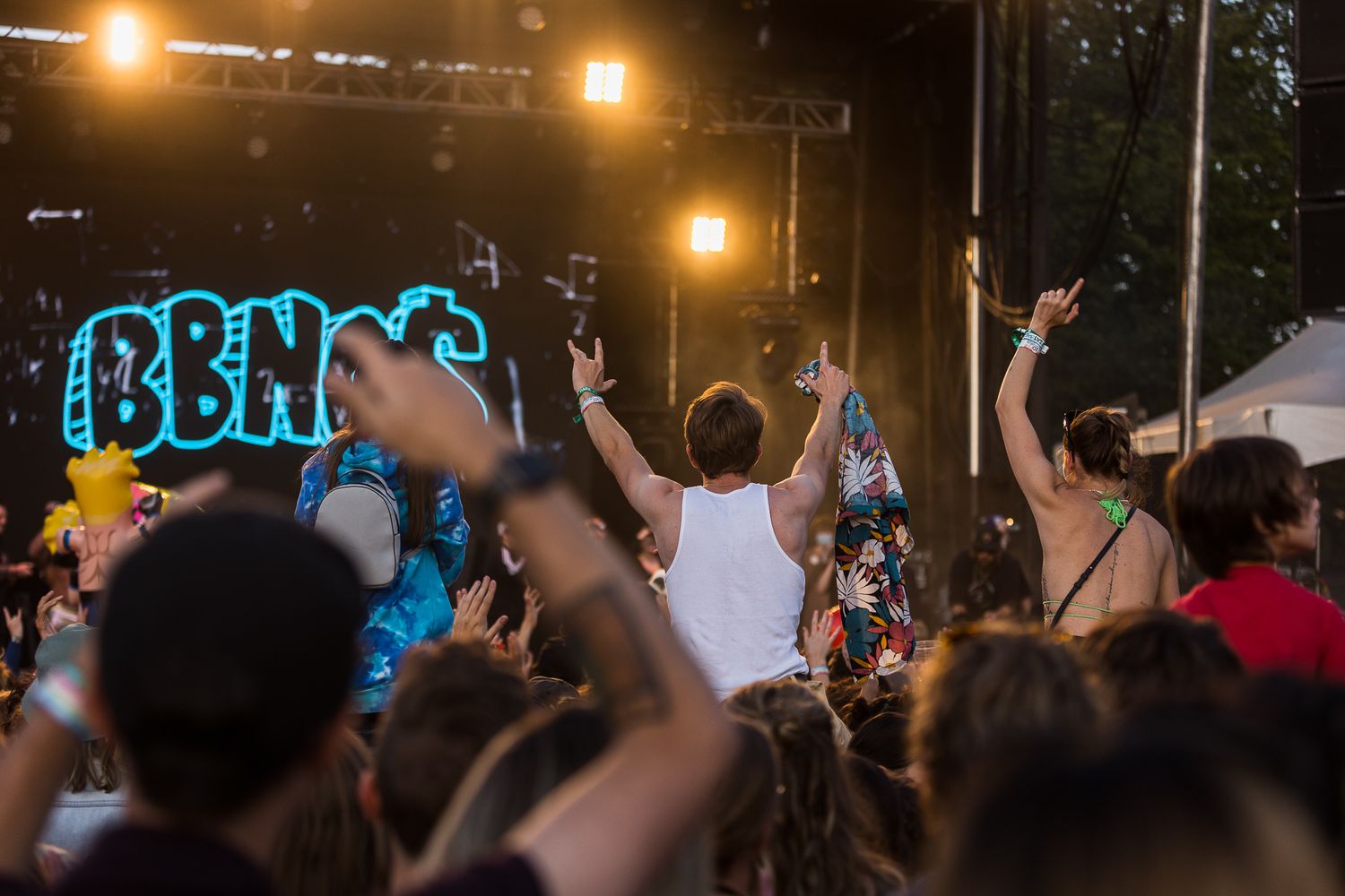 Concert crowd with raised hands watching performance at outdoor music festival with illuminated stage at night.