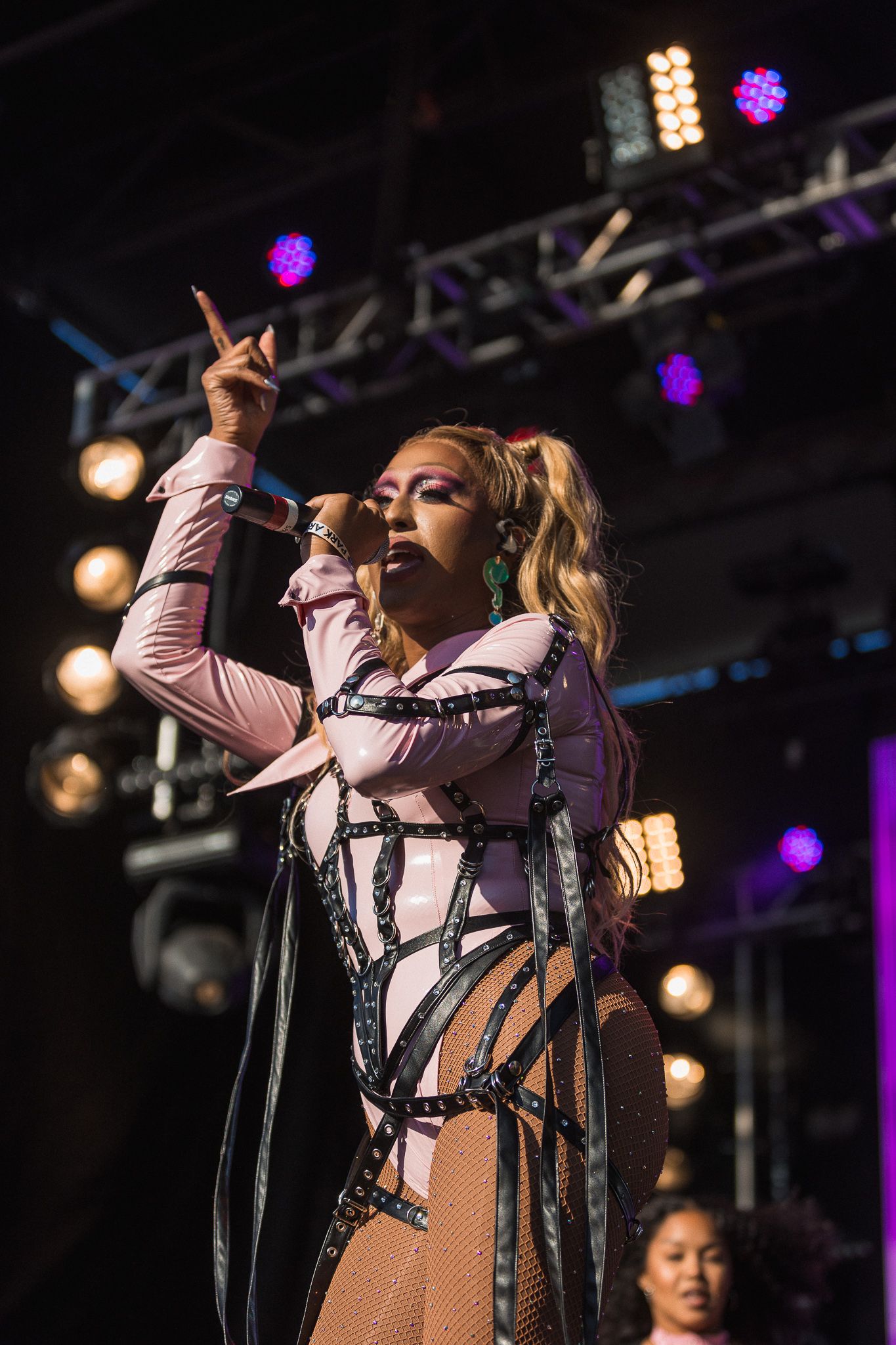A performer in a pink and white outfit sings on stage with dramatic lighting and raised hand gesture.