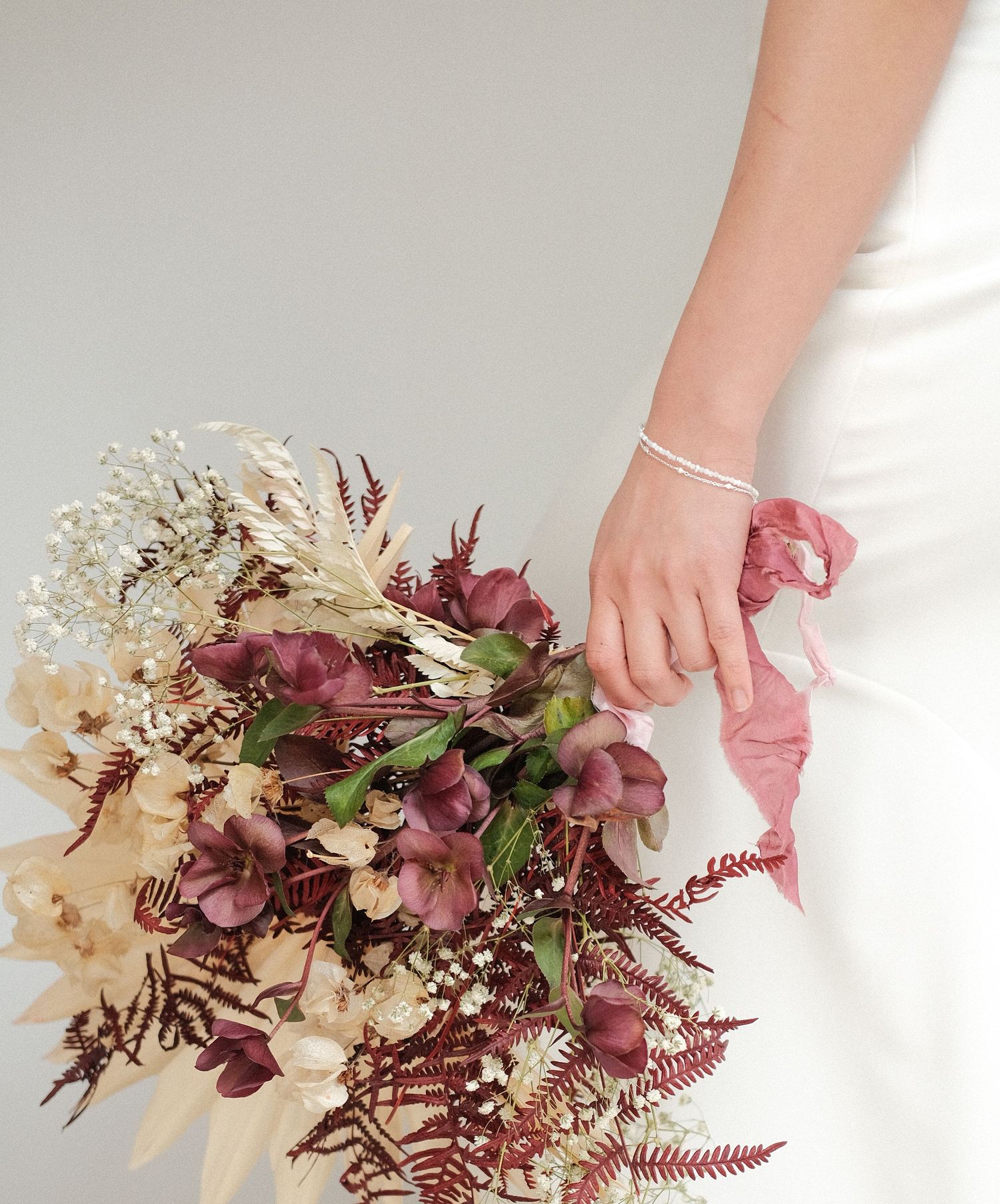 A delicate bridal bouquet with burgundy and white dried flowers, featuring baby's breath and ferns against a white dress.