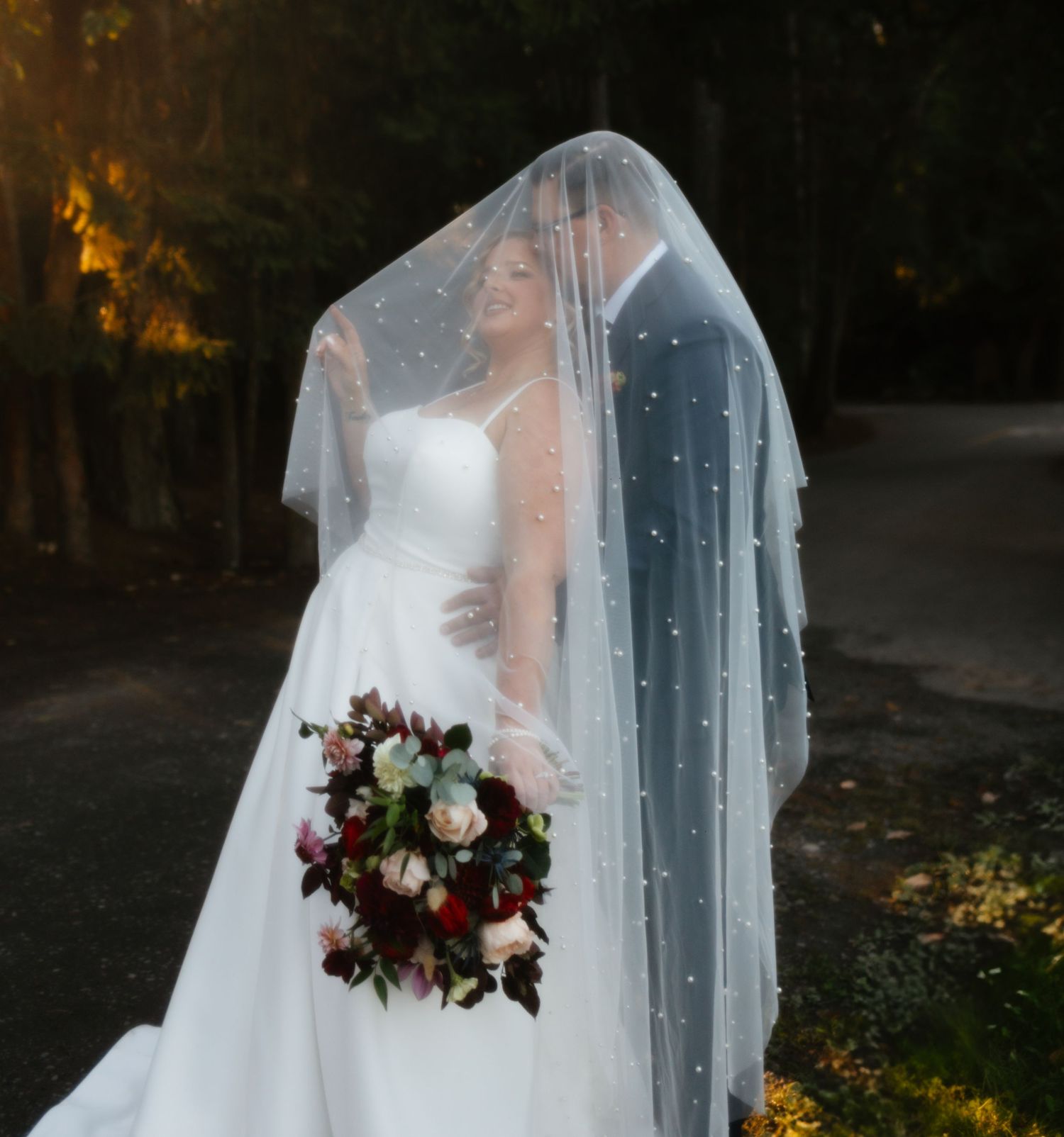 A bride holds a deep burgundy and cream floral bouquet while wearing a flowing white gown and sheer veil in an autumn setting.