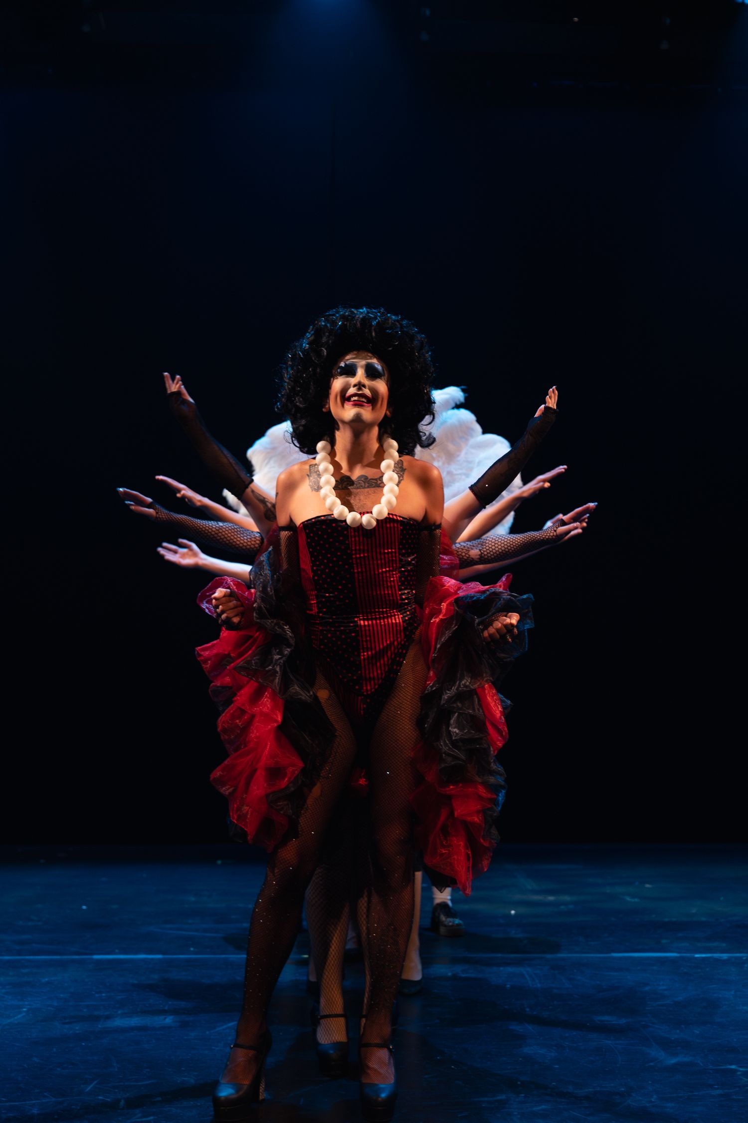 Four performers in glamorous red showgirl costumes with feathers dance on a dark stage under blue spotlight.