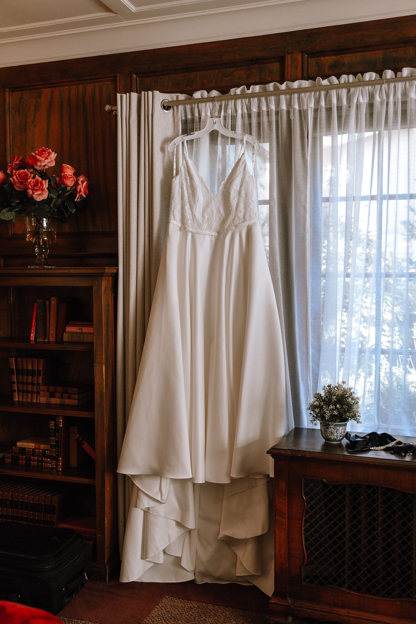 An elegant white wedding dress hangs by a sunny window in a wood-paneled room with bookshelves and floral arrangements.