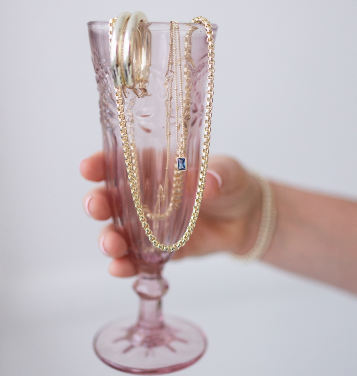 Delicate gold chains and necklaces draped elegantly over a pink vintage-style champagne glass.