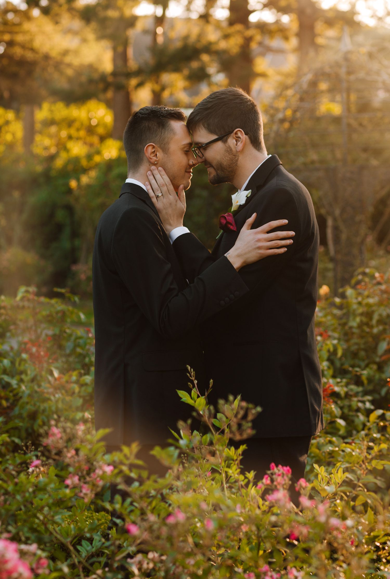 Two grooms share an intimate moment in a garden at sunset surrounded by flowers.