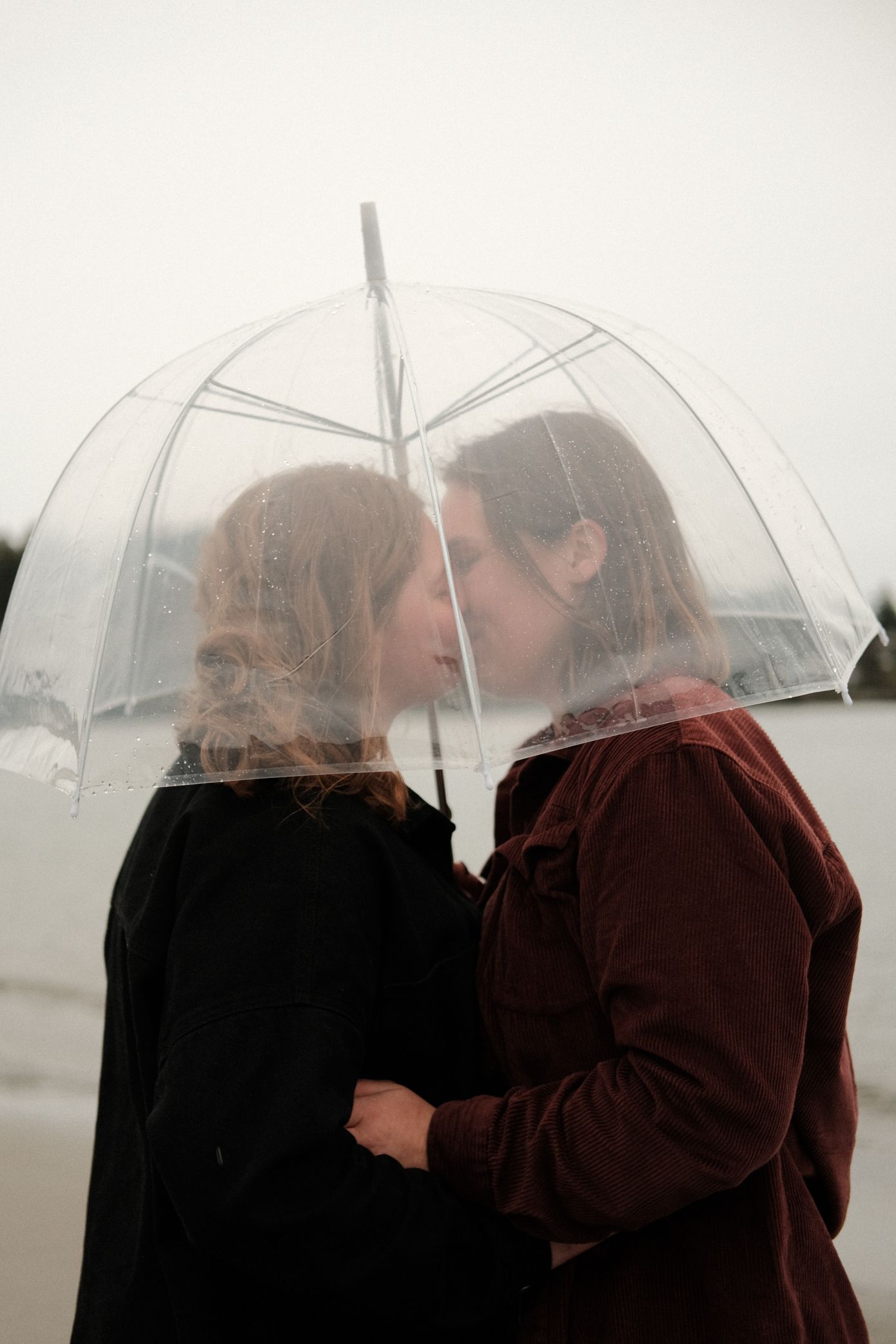 A couple shares an intimate moment under a clear umbrella on a misty day, dressed in dark winter coats.