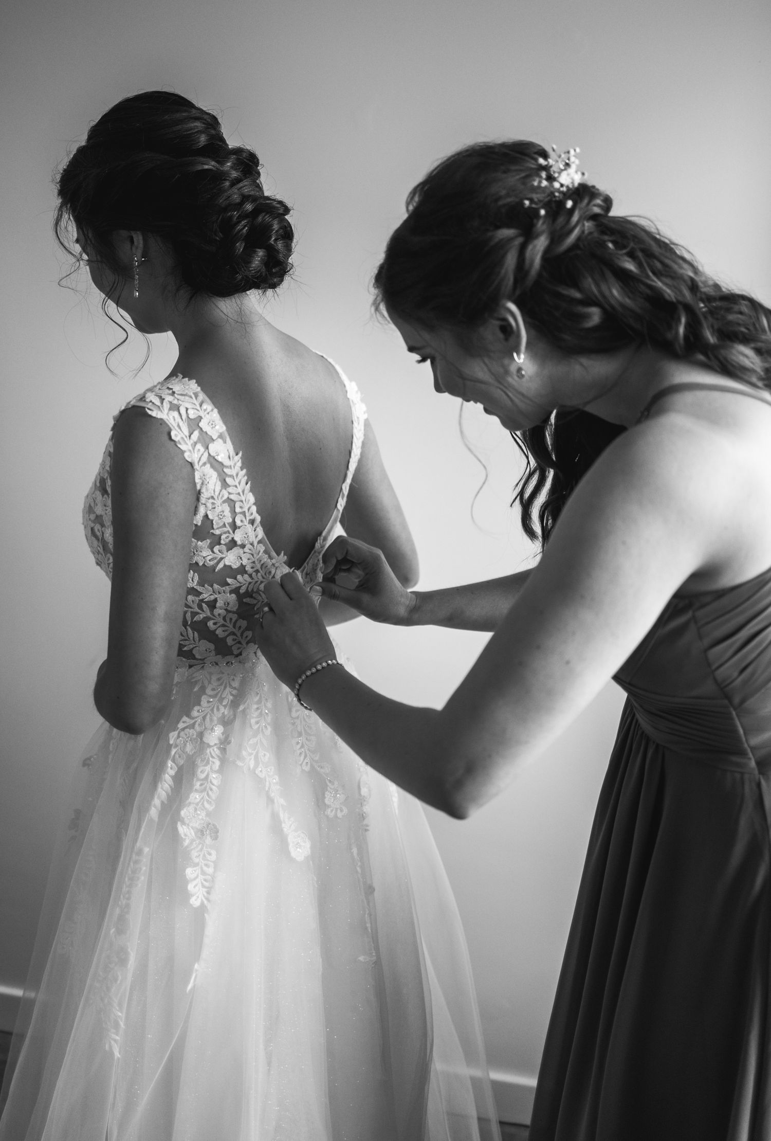 A tender moment of getting dressed for a wedding, showing the intricate lace details of a white bridal gown being fastened.