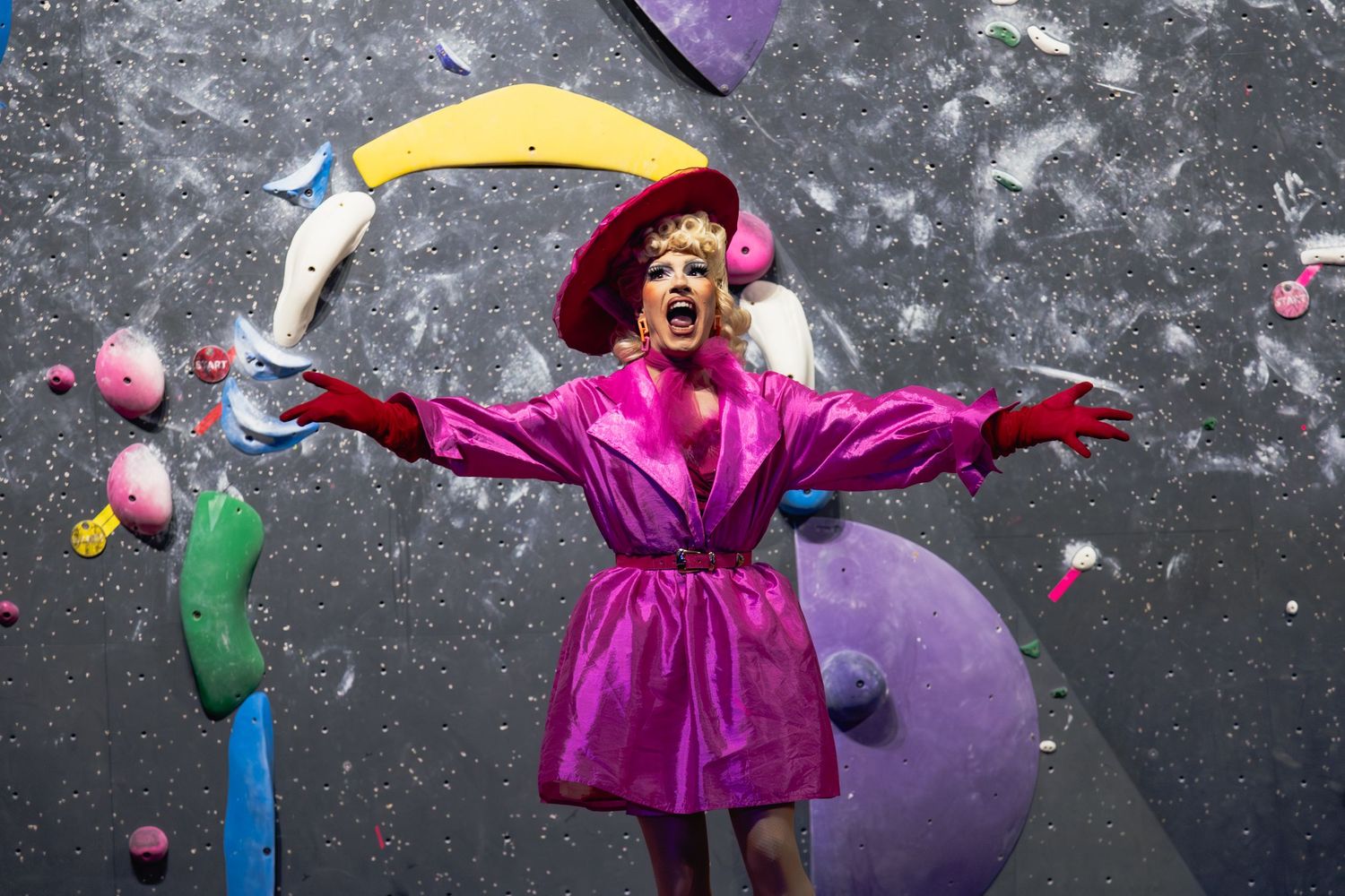 A performer in a bright pink robe poses dramatically against a dark wall with colorful balloon decorations.